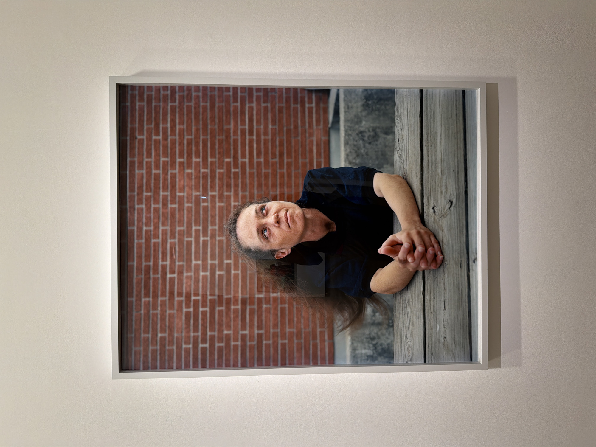 A framed photograph hangs vertically on a plain, light beige wall. The frame is simple and light grey or white. The image depicted within the frame is rotated 90 degrees clockwise from its natural orientation, showing a woman with her head positioned towards the left side of the frame and her body extending to the right.
The woman has long, dark brown hair, fair skin with freckles, and is looking upwards with a contemplative or pensive expression. She is wearing a dark, short-sleeved top. Her hands are clasped together and resting on a textured, weathered wooden surface, which appears to be a fence, railing, or table. Behind her, to the left (which would be above her in a corrected orientation), is a distinct red brick wall with a repeating pattern. To the right of the wooden surface (below her arms), the background is darker and less defined.
The scene within the photograph suggests an outdoor setting, indicated by the brick wall and the weathered wood. Natural light illuminates the subject, implying it was taken during the daytime. There is no visible text in the image. The overall media file captures an art piece displayed indoors.