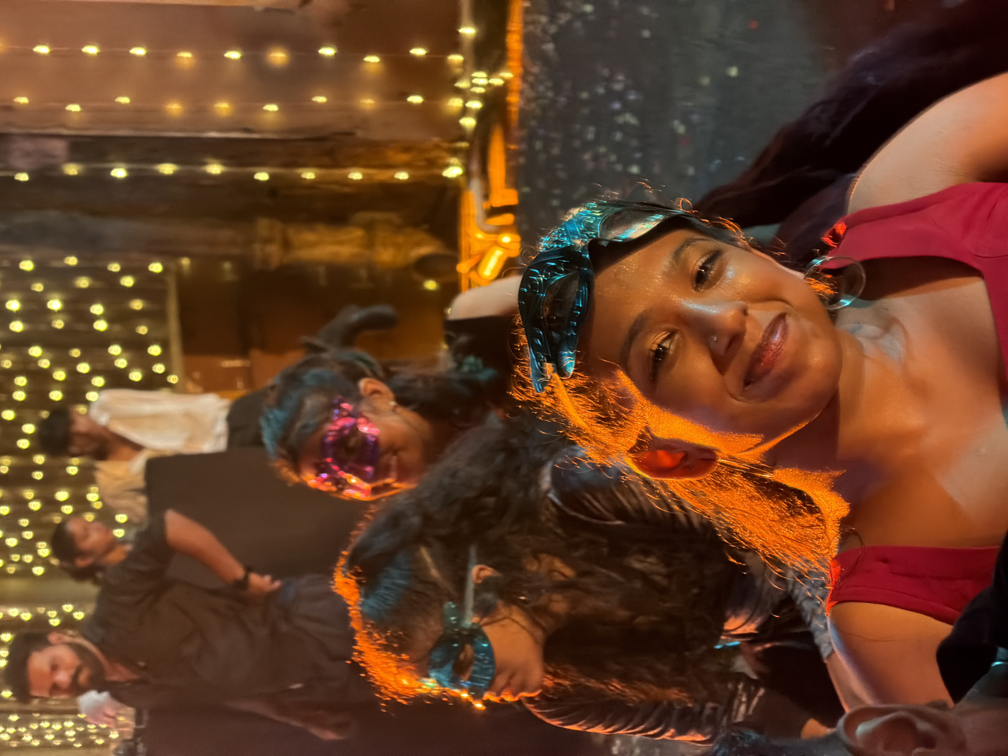 The image captures a vibrant and festive night gathering in Kalamassery, India. The primary subject, a woman, is positioned prominently on the right side of the frame, smiling and looking directly at the viewer. She has dark skin, dark curly hair, and wears a red top along with a distinct dark blue patterned headband and large hoop earrings. Her face is warmly illuminated by an orange glow, suggesting an artificial light source.
Behind her and extending to the left, several other people are visible, all contributing to the lively atmosphere. One individual, slightly behind the main subject, wears a sparkly purple masquerade mask and dark clothing, with dark, curly hair. Another person in the mid-ground also appears to be wearing a blue or teal masquerade mask. Further in the background, two men are partially visible; one with a beard wears a dark shirt, and another person in a white top is seen in the upper left background.
The setting is characterized by numerous rows of warm, yellow string lights that are strung across what appears to be wooden beams or structures. These lights create a soft, bokeh-like effect, indicating a celebratory event at night or in a dimly lit indoor space. The presence of masquerade masks strongly suggests a themed party, a carnival, or a festive social gathering. The overall mood conveyed is cheerful and engaging, with the main subject's smile reflecting enjoyment of the event. No visible text is present in the image.