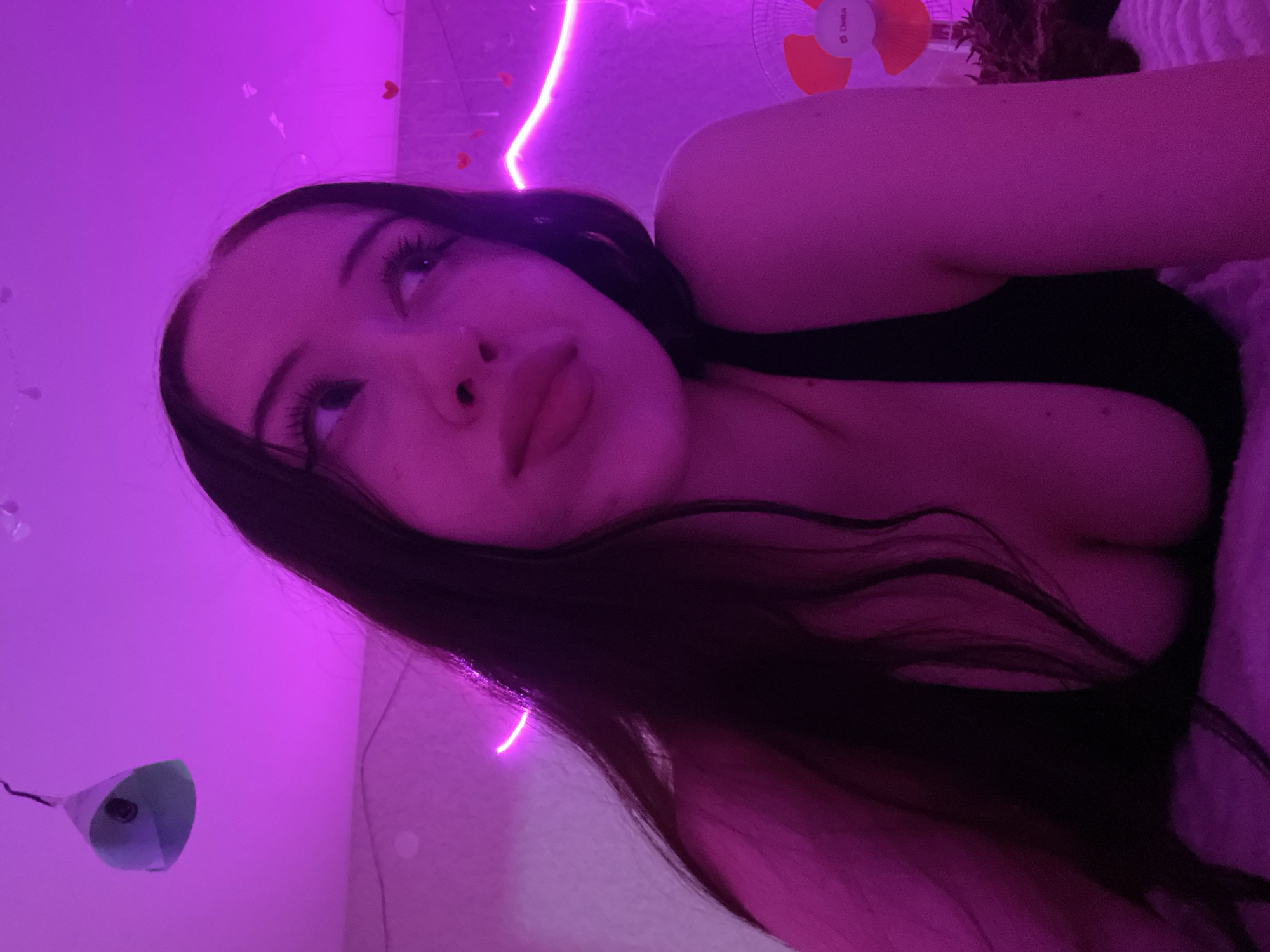 A young woman with long, dark hair is lying down in a room illuminated by purple neon lights. She is wearing a black top. Her eyes are looking upwards and to the side. In the background, there are small red heart decorations, a string of neon lights, a ceiling decoration resembling a paper lantern, and a small fan. The overall impression is one of a casual, possibly indoor, setting. The mention of Votkinsk, Russia, is not supported by any visual cues in the image.