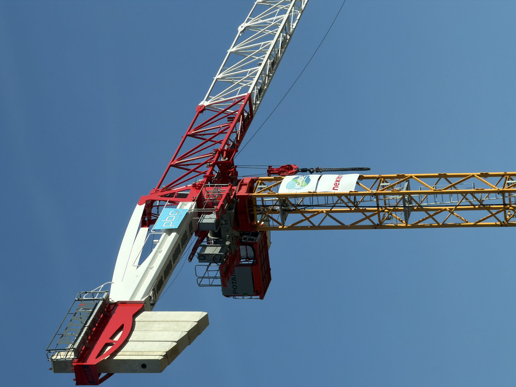 This image shows a construction crane against a clear blue sky. The crane is red and white, with a yellow lattice tower. There are several logos visible on the crane, including "POTAIN," "PBC," "ICADE," and "nexity." The crane appears to be in operation, with a hook and chain suspended from the jib. The scene is located in the city of Bron, France.