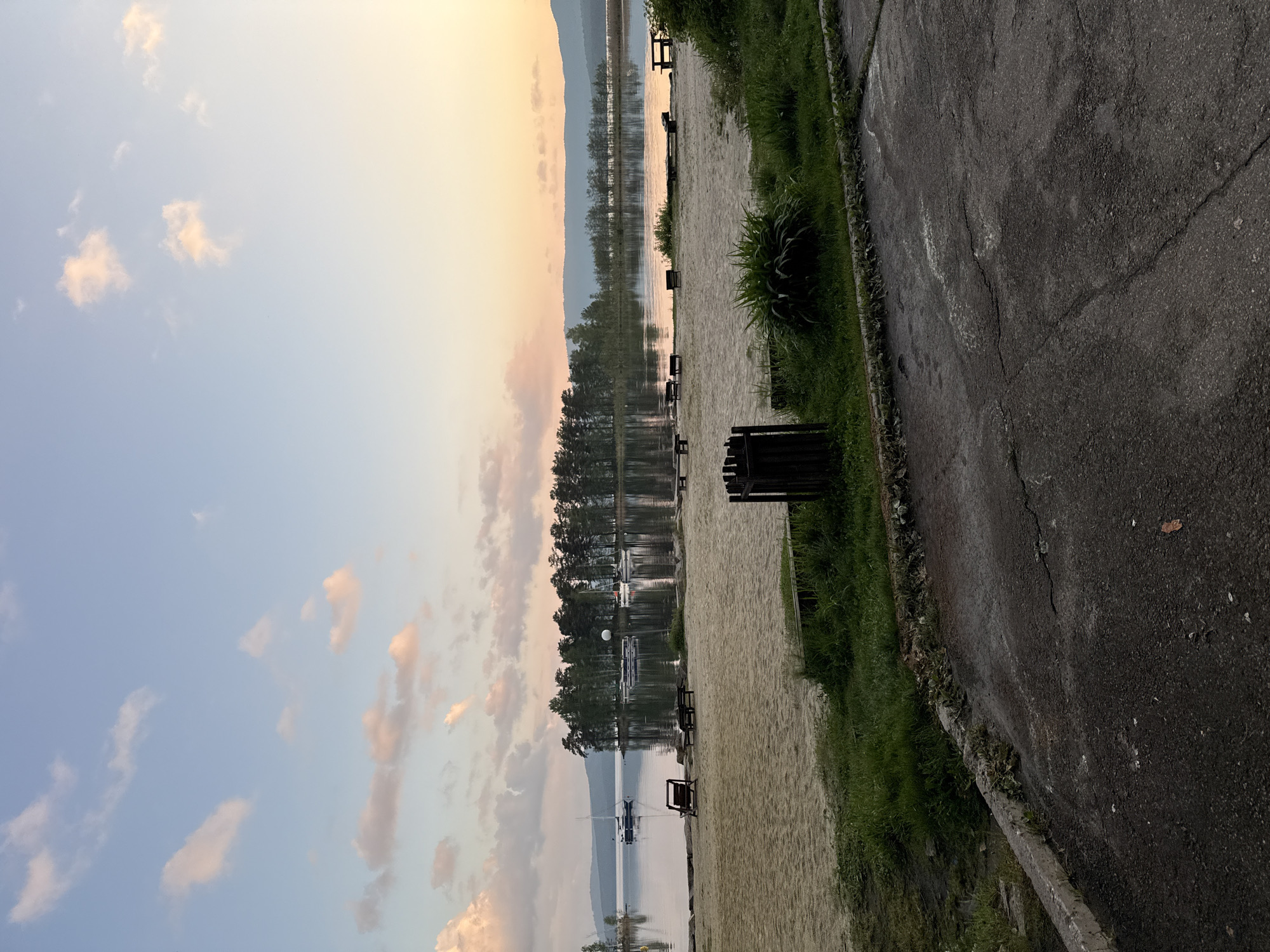 The image shows a lakeside view in Zolotoi Plyazh, Russia. A body of water reflects the sky and surrounding trees, suggesting calm conditions. Benches line the edge of a sandy area beside the lake. A paved walkway runs parallel to the sandy area, marked by cracks and edged with grass. In the distance, a mountainous terrain is visible under a sky tinted with colors of a setting or rising sun. There are boats docked on the water. No people are visible in the frame.