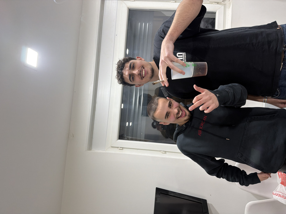 Two young adult males are depicted indoors, posing for a photograph. The subject on the left has short, dark buzz-cut hair and a goatee, wearing a black hooded sweatshirt with "AIR JORDAN" in red text on the chest. He smiles broadly with an open mouth and extends his right hand, forming a gesture with his index and pinky fingers raised. The subject on the right has curly dark hair and a mustache, wearing a black t-shirt with a discernible white graphic (likely a brand logo, partially obscured). He smiles while holding a clear plastic cup containing a dark liquid in his left hand and a lit, hand-rolled cigarette, emitting smoke, between the index and middle fingers of his right hand. Both individuals face the camera. The background consists of plain white walls, a large white-framed window reflecting interior elements including a partial figure holding a device, and a white ceiling with a single square recessed light fixture. A black television screen is partially visible to the far left, along with a white plastic shopping bag displaying the "CONTINENTE" logo and a white chair. The scene is illuminated by artificial indoor lighting. This scene is located within the city of Bragança, Portugal.