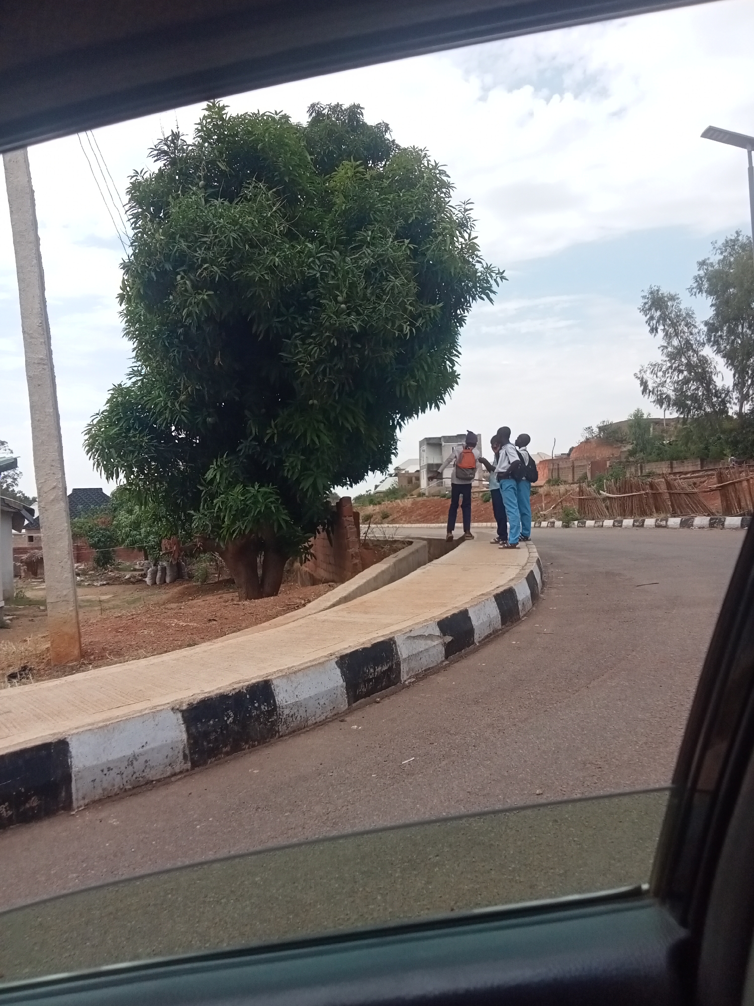 The image depicts a street scene in Jos, Nigeria. A large, leafy tree dominates the left side of the frame, adjacent to a concrete utility pole. A raised concrete curb with alternating black and white painted blocks borders the roadway. Three or four young people, possibly students in uniforms consisting of blue pants and white shirts with backpacks, stand on the curb. The road curves to the right, with fencing and some houses visible in the background on a slightly elevated area. The image is taken from inside a vehicle, indicated by the car window in the foreground. The sky is overcast.