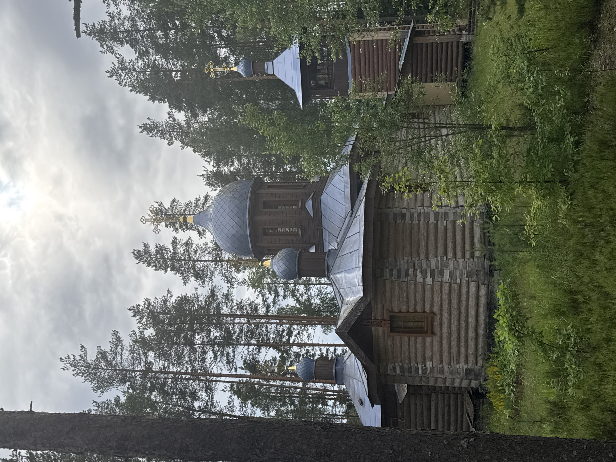 The image shows a wooden Orthodox church in Nenyamyaki, Russia. The church has several domes topped with crosses and a bell tower. It is surrounded by tall pine trees and lush green vegetation. The sky is overcast with some sunlight breaking through the clouds. The church is constructed from logs, with stone accents around the windows. The bell tower has exposed wooden beams and a metal roof. There are no people visible in the image.