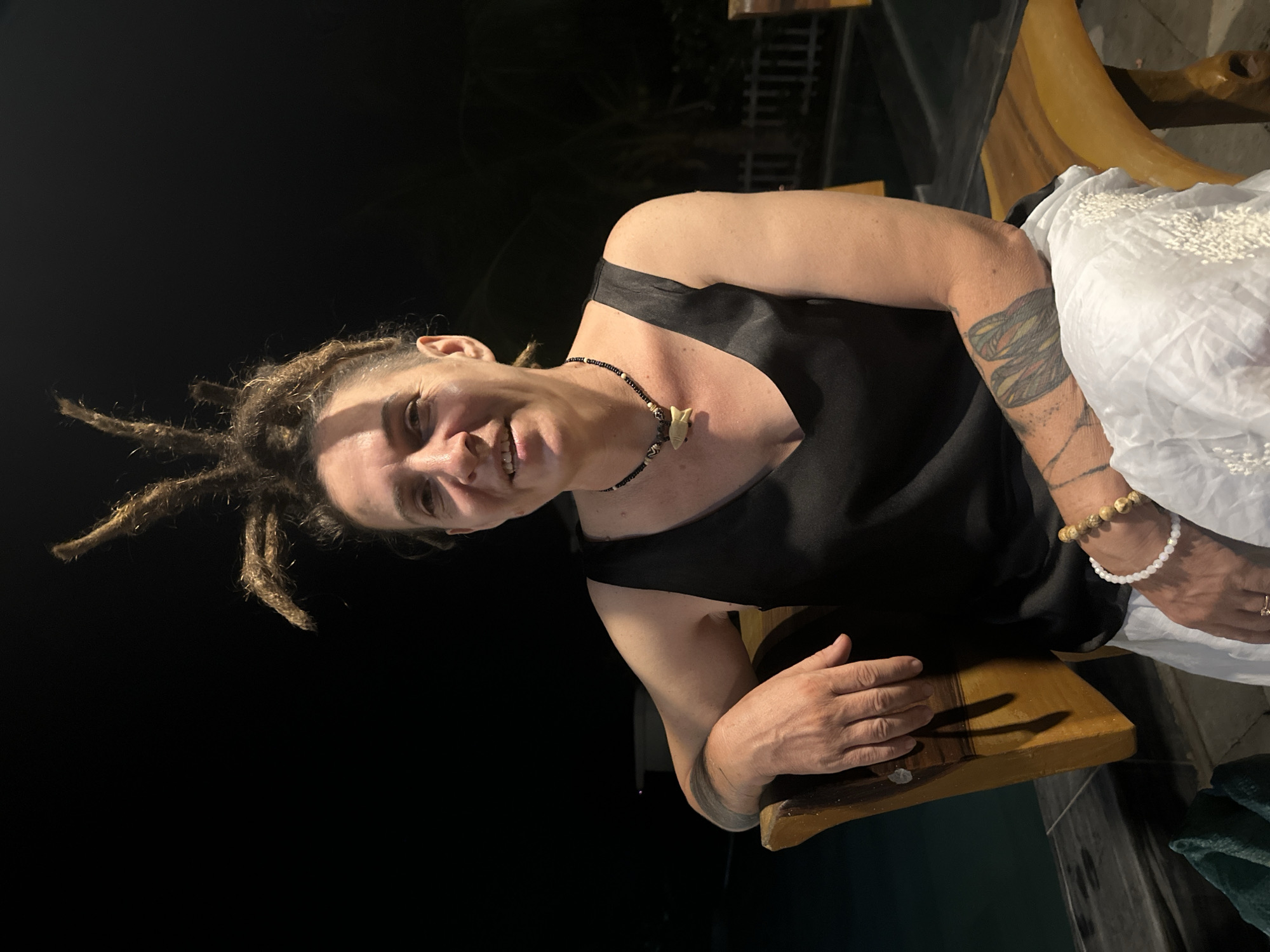 The image is a medium shot of a woman with dreadlocks sitting outside at night. She is light-skinned, wearing a black tank top and a necklace with a light-colored pendant. She has tattoos on both arms and is wearing bracelets on her right wrist. Her left hand rests on a wooden surface. A white garment is draped over her right side. The background is dark and suggests an outdoor setting. The image was taken in the city of Mũi Né, Vietnam.
