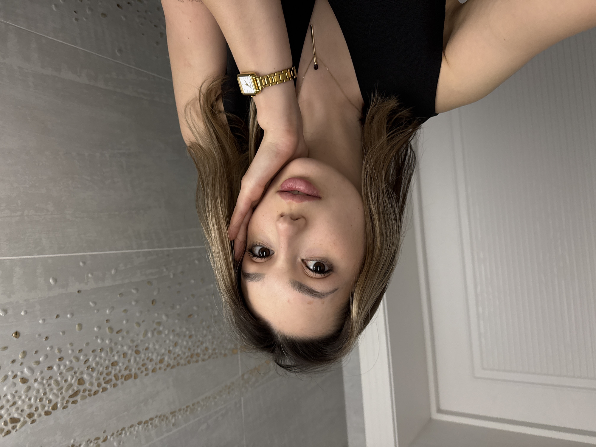 A young woman with shoulder-length brown hair poses for a selfie in what appears to be a bathroom. She is wearing a black sleeveless top, a gold watch on her left wrist, and a delicate gold necklace. Her left hand is resting against her face. The background consists of a wall with gray tiles on the left and a white door on the right. The city and specific location are unknown.