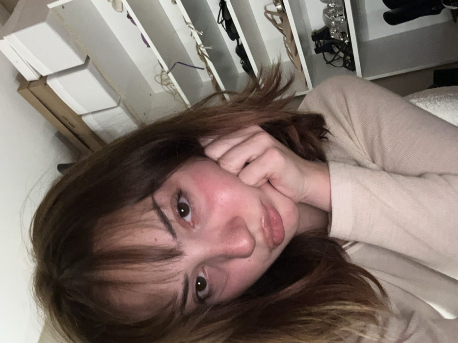 A woman poses for a selfie. She rests her cheek on her hand. Her dark hair falls around her face, framing it with bangs. She has light skin. Behind her is a white closet with shelves holding boxes and shoes. She wears a light-colored shirt. The photo is taken at an angle from above.