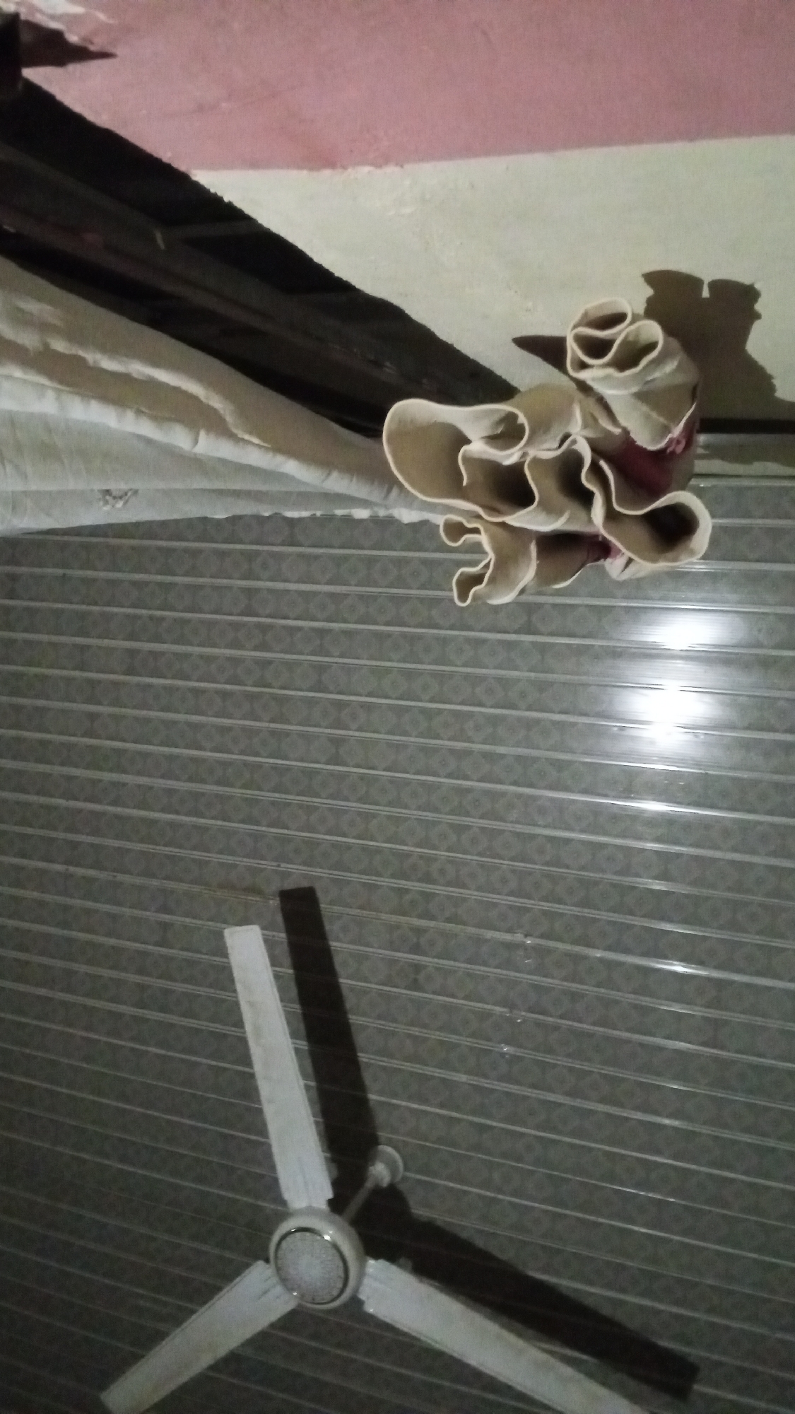 The image depicts an interior scene, likely a room, with a ceiling fan and what appears to be a rolled-up fabric or tarp hanging from the ceiling. The ceiling has a pink and white painted section above a dark wooden beam. Below this, a beige, tightly rolled item with distinctive folds is suspended. The lower part of the image shows a horizontally striped patterned wall, characteristic of some interior paneling. A white ceiling fan with three blades is visible in the bottom portion of the frame, casting a dark shadow on the patterned wall. The lighting suggests it is nighttime or a dimly lit interior. The scene appears to be an indoor domestic setting, potentially in Jalingo, Nigeria, given the provided context, although specific Jalingo landmarks are not identifiable in the image.