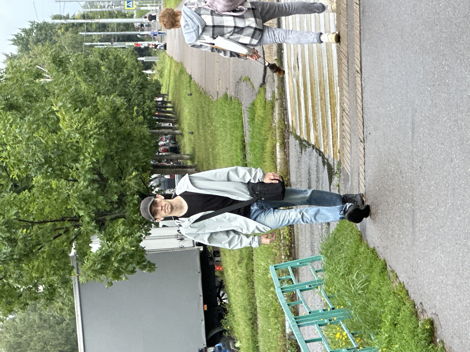 A man wearing a light gray jacket, black t-shirt, blue jeans, black baseball cap, and a black crossbody bag walks on a paved path towards the camera. He is on the left side of the image. A person in a gray and white checkered jacket, gray pants, and a light pink backpack walks away from the camera on the right side of the image, holding a dog on a leash. The dog is brown and appears to be a medium-sized breed. Both individuals are walking on a wet, grey paved path adjacent to a grassy area with a green metal fence. The path leads to a pedestrian crossing marked with yellow and white stripes. Trees line the background on the left side, and utility poles and streetlights are visible on the right. In the far left background, a large grey truck is parked. The scene appears to be in an urban or suburban environment, possibly in Podolsk, Russia, given the context. The overall lighting suggests an overcast day.