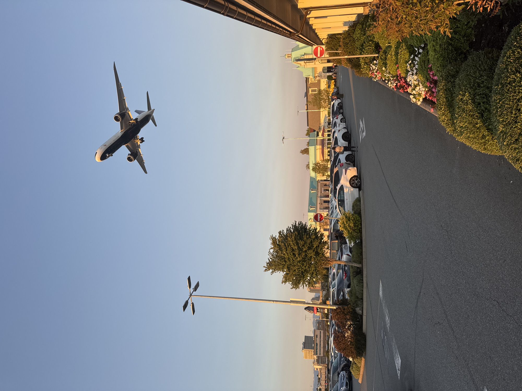 An aerial view captures a commercial district in Richmond, Canada, under a clear sky during the golden hour. A passenger jet is descending for landing, its landing gear deployed, in the upper portion of the frame. In the middle ground, a large parking lot is filled with numerous cars. Several buildings with light-colored facades and green roofs are visible in the background, suggesting a retail or outlet mall environment. A tall lamppost with four lamps stands prominently on the left side of the parking lot, and a tree with dense green foliage is situated near it. A red and white circular sign with a horizontal red bar, indicating "Do Not Enter," is placed next to the entrance of a walkway. The foreground features a paved road with visible lane markings, and on the right side, meticulously manicured bushes and flowerbeds add a touch of natural beauty to the scene. The warm sunlight casts long shadows, highlighting the textures of the asphalt and the foliage, and creating a serene atmosphere.