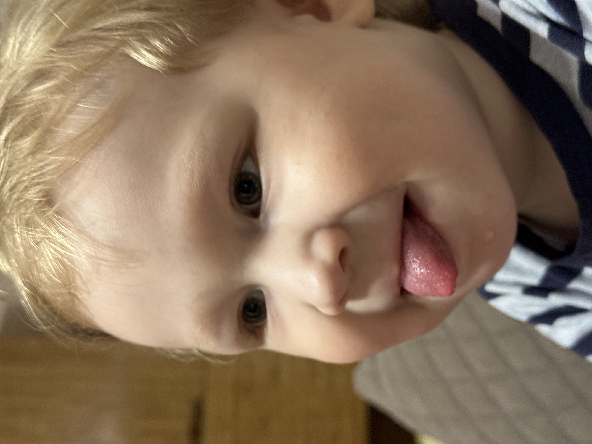 A young child with fair skin and light blonde hair is sticking out their tongue. The child's eyes are brown. They are wearing a dark blue and white striped shirt. The background includes a wooden floor and a light-colored sofa or bedding. There is no evidence to suggest the location is Votkinsk, Russia.