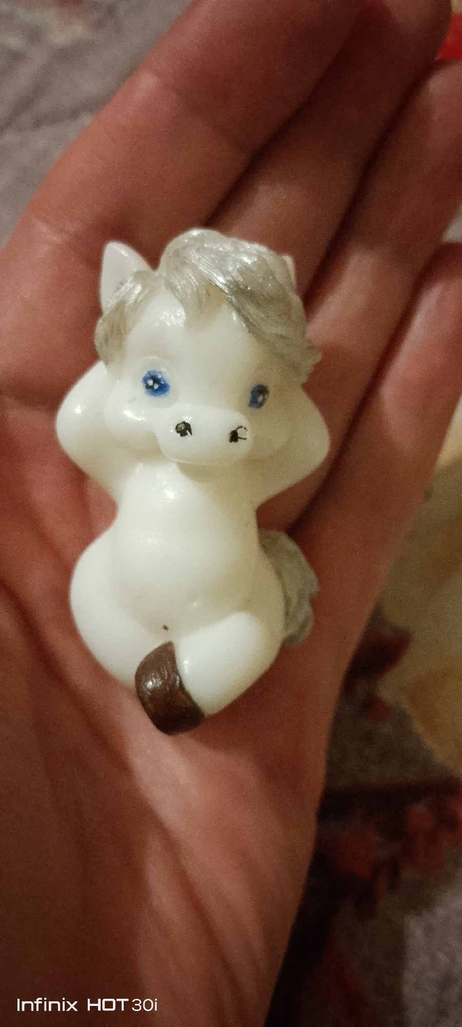 A white, plastic figurine of a horse is held in a person's hand. The horse has blue eyes and a gray mane. Its body is plump and it is sitting with its legs tucked underneath. A brown hoof is visible at the bottom of the figurine. The background is out of focus, but appears to be a textured surface, possibly fabric. The timestamp "Infinix HOT 30i" is visible in the bottom left corner of the image. The context of Kedrovy, Russia is provided.