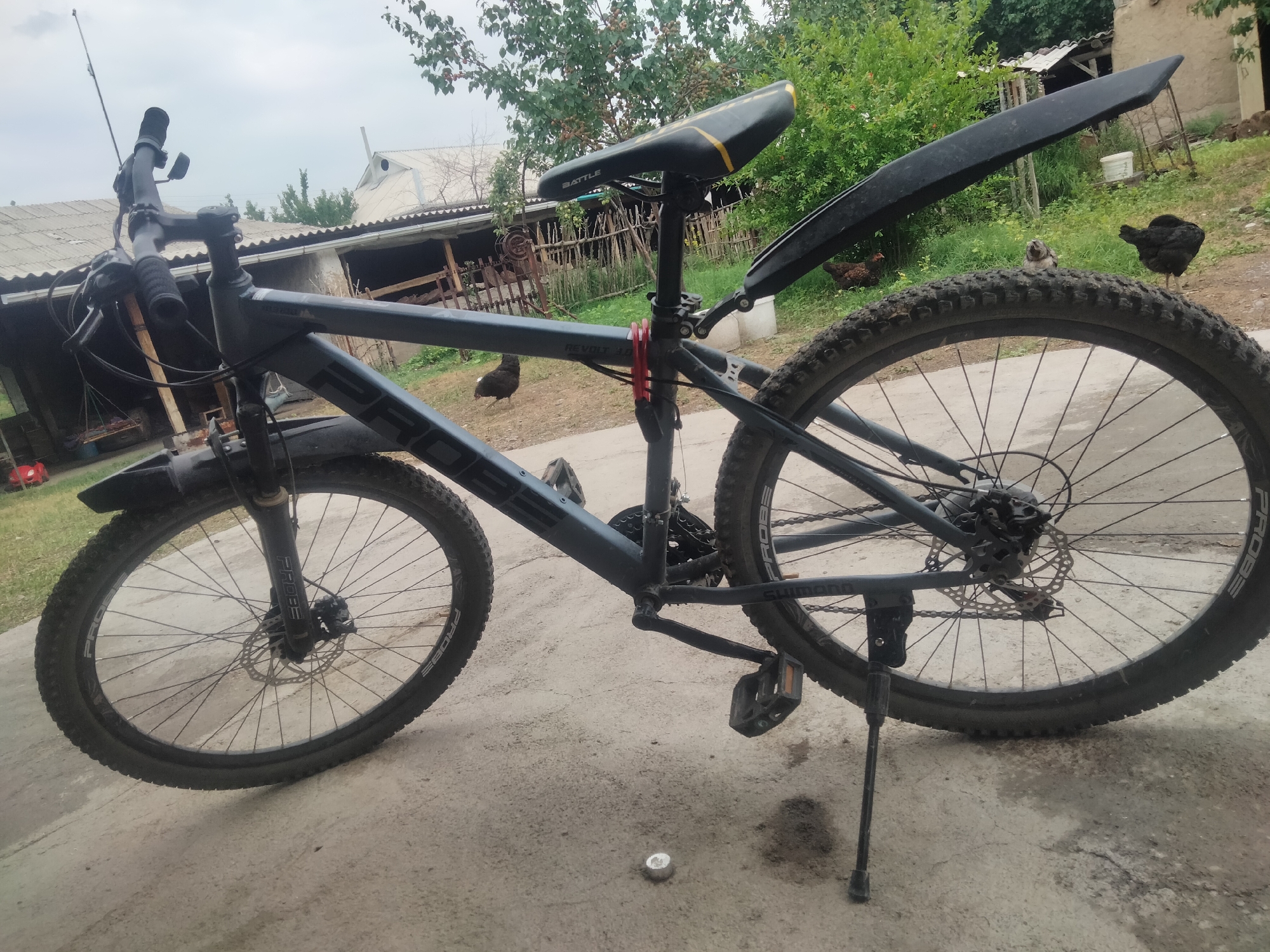 A dark grey mountain bike is parked on a concrete surface in an outdoor environment. The bike has a black saddle with yellow accents, black fenders, and knobby tires. The rear fender is extended upwards and backwards. A kickstand is deployed, supporting the bike. In the background, there are several chickens, some buildings with corrugated metal roofs, a wooden fence, and trees. The sky is overcast. The scene appears to be in a rural or semi-rural setting, likely a yard or courtyard. The location is identified as Baş-Bulak, Kyrgyzstan.