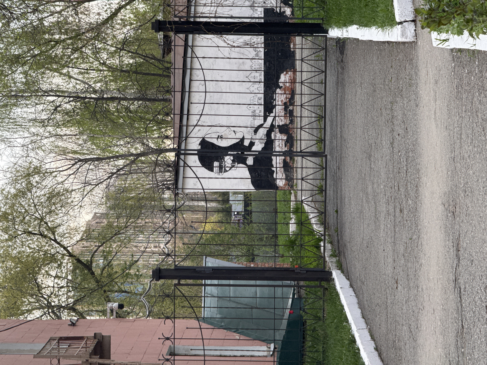 The image shows a black metal gate in Izhevsk, Russia, with a large mural visible through it. The mural features a woman's face in black and white, stylized with a city skyline above her head. The mural appears to be painted on a wall behind the gate, possibly part of a building. The gate is closed, and a paved pathway leads up to it. There is some green grass and foliage visible on either side of the pathway and behind the gate. The buildings surrounding the area appear to be a mix of residential and possibly commercial structures. A security camera is visible on the lower left of the gate.