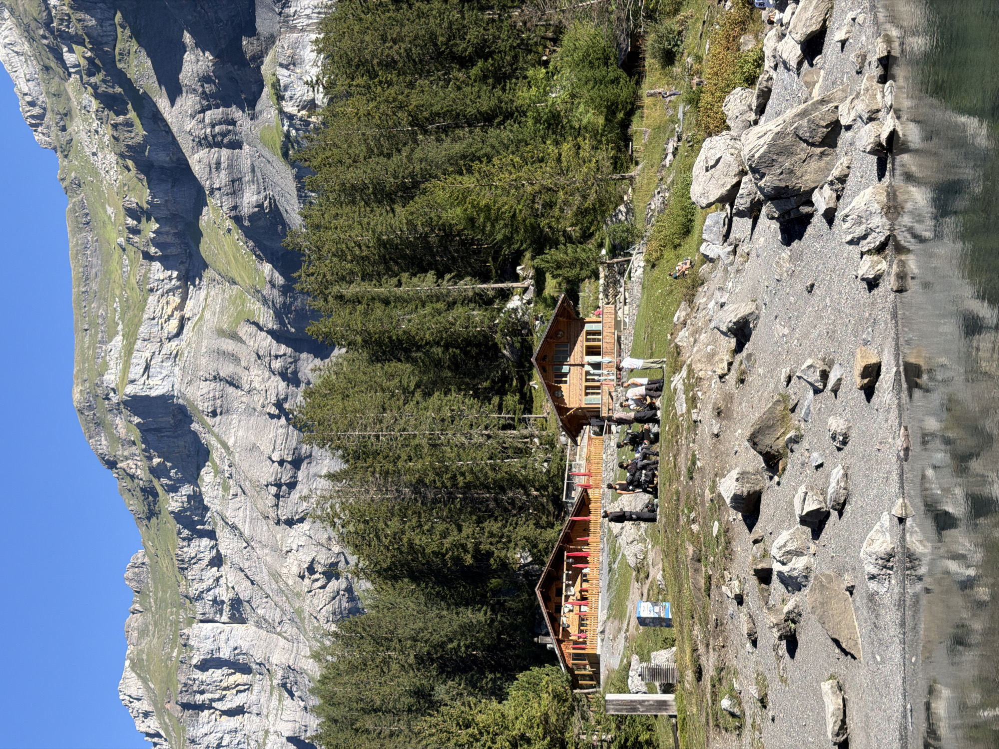 The image depicts a serene mountain landscape with two traditional wooden chalets nestled amidst a dense pine forest, with a rocky shore in the foreground leading to a calm body of water. The chalets, one larger and more prominent than the other, are characterized by their wooden construction, pitched roofs, and extensive outdoor seating areas with red umbrellas. A group of people are gathered near the chalets, some seated and others standing, engaged in conversations or relaxing. The background is dominated by a majestic, rugged mountain range with layered rock formations and patches of green vegetation under a clear blue sky. The water in the foreground reflects the mountain scenery and the sky, adding to the tranquility of the scene. This scene is located at Oeschinensee, a high-alpine lake in the Swiss Alps near Kandersteg, Switzerland, renowned for its breathtaking natural beauty and recreational activities.
