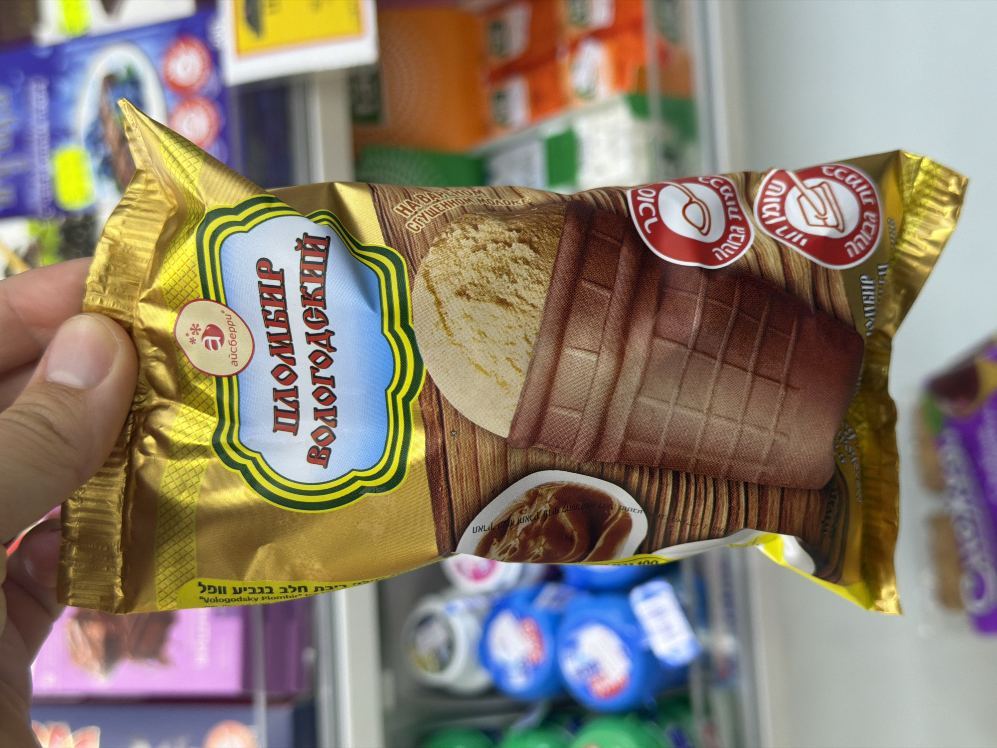 A hand is holding a gold-colored package of "Vologodsky Plombir" ice cream, featuring an image of a waffle cone with chocolate and ice cream. The package has inscriptions in Russian and Hebrew. The image suggests the ice cream is for sale at a location in Tel Aviv, Israel, inside a freezer stocked with other ice cream and snack products.