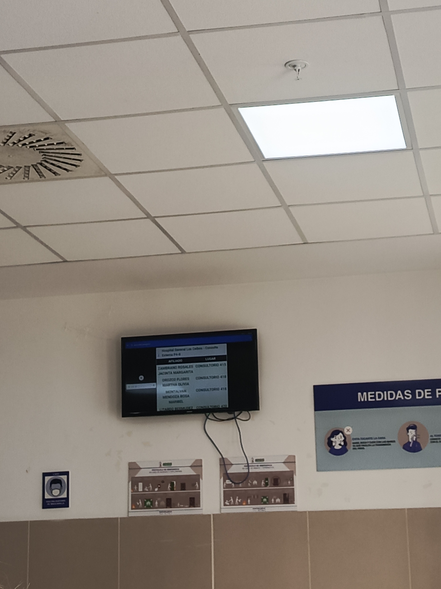 This media file depicts an indoor scene within a medical facility, likely a hospital or clinic, in Guayaquil, Ecuador. The primary focus is a digital display screen mounted on a white wall, showing a list of names and associated consultation room numbers. The screen lists names such as "ZAMBRANO ROSALES," "JACINTA MARGARITA," "OROZCO FLORES," "MARTHA OLIVIA," "MONTALVAN," "MENDOZA ROSA," and "MARIBEL," each with a corresponding "CONSULTORIO" (consultation room) number, for instance, "415" and "418." The header text above this list indicates "Hospital Guayaquil" and "Externa P4-0."
To the right of the digital display, a dark blue sign with white text reads "MEDIDAS DE F," suggesting health or safety measures. Below this sign, there are two circular illustrations, one showing a person wearing a mask with an "X" over it, and another showing a person coughing.
To the left of the digital display, there's a smaller blue square sign with a white icon of a person wearing a mask. Below the digital display and to the left, there are two framed posters on the wall. One poster appears to be an informational graphic with various illustrations, possibly depicting safety protocols or facility information. The ceiling is visible at the top of the image, with visible light panels and a circular air vent, typical of institutional settings. The lower portion of the image shows a wall with a tiled or panel finish in a neutral tone. The overall impression is of a functional, public space within a healthcare institution.