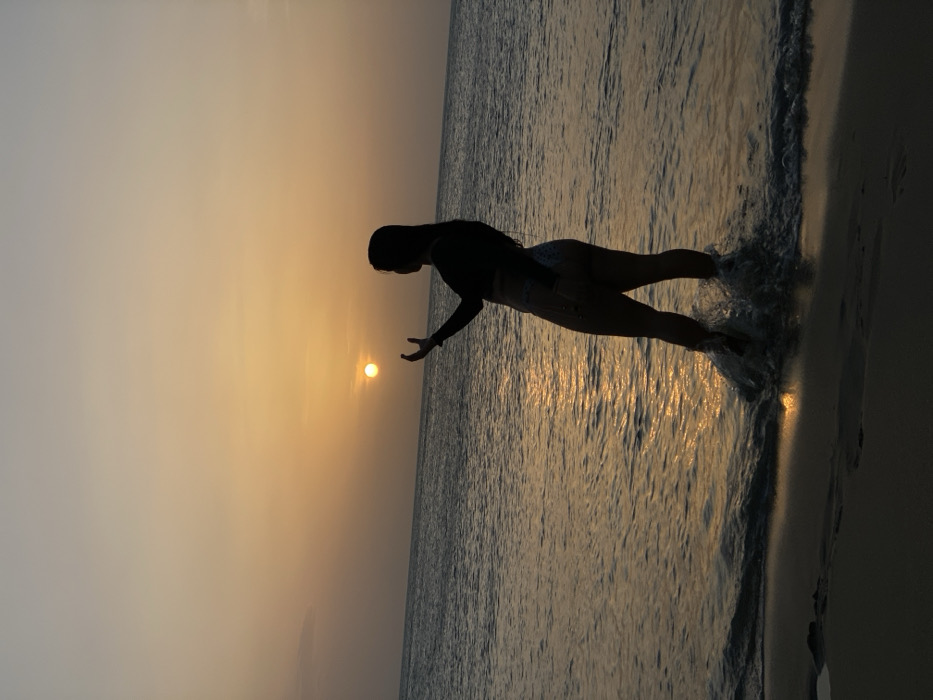 The image depicts a silhouetted individual, likely female, standing in the shallow waters of the ocean at sunset in Juan Griego, Venezuela. The subject is oriented towards the left side of the frame, with her head slightly turned to her right, facing the setting sun. She wears a dark, long-sleeved top and light-colored, possibly patterned, bikini bottoms. Her right arm is raised, with an open hand positioned to optically "hold" the bright, circular disc of the sun visible on the horizon. Waves are actively splashing around her lower legs and feet, creating white foam. The sky transitions from a vibrant orange-yellow at the horizon to a lighter, paler yellow higher up. The setting sun casts a reflective orange-yellow path across the surface of the ocean water, extending towards the viewer. The immediate foreground consists of dark, wet sand.