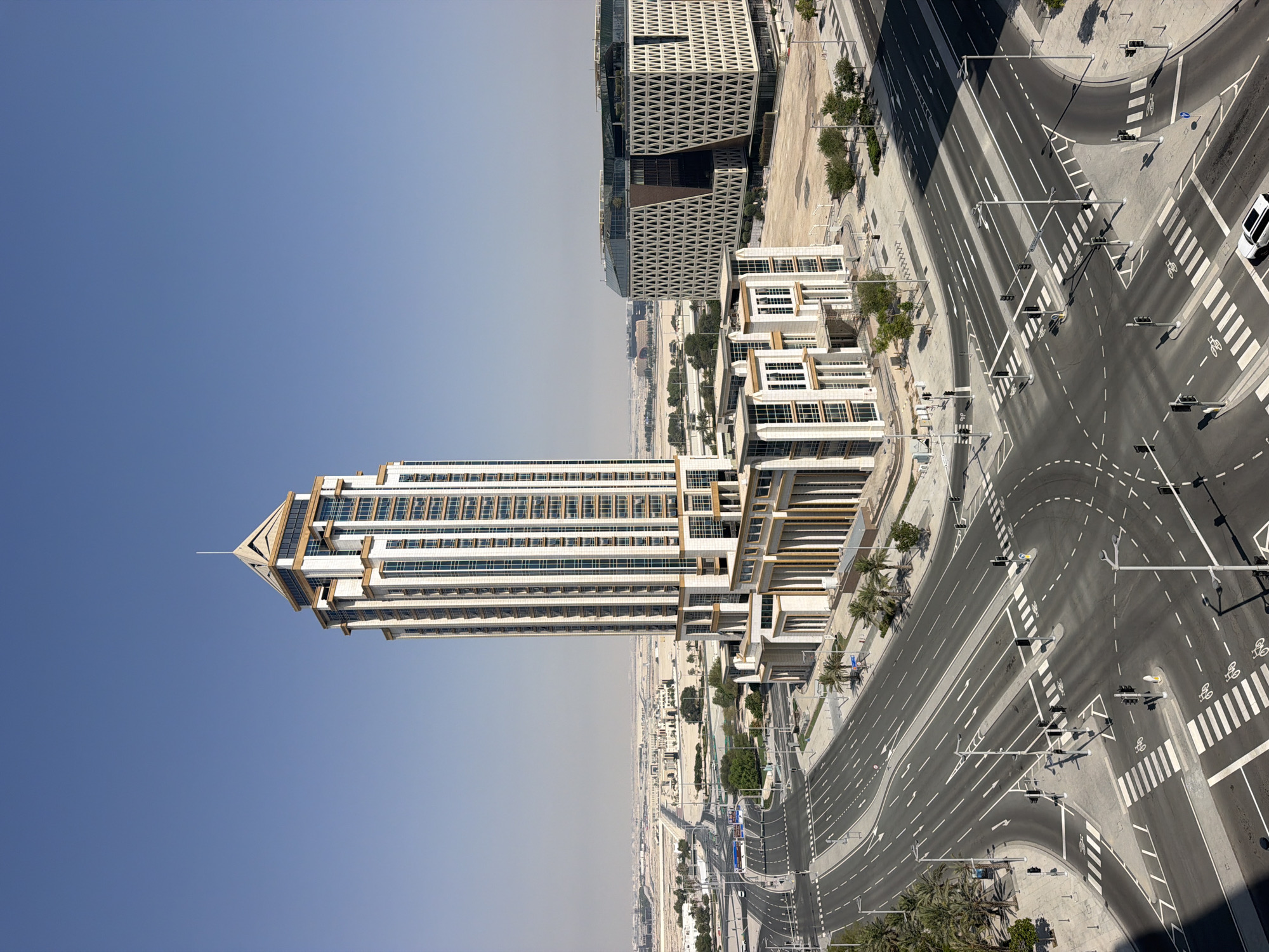 An aerial view shows a modern cityscape under a clear blue sky. Dominating the foreground is a tall, white skyscraper with gold accents and a stepped, pyramidal top. To its right, a lower, modern building features a distinctive white, triangular geometric facade. In front of these buildings, a wide, multi-lane intersection with clear road markings, pedestrian crosswalks, and traffic signals is visible. Palm trees and other sparse vegetation line the roads and sidewalks. In the distance, a flat, arid landscape stretches to the horizon, dotted with more contemporary structures. The scene depicts an urban environment, likely in a warm climate, with a focus on modern architecture and infrastructure.