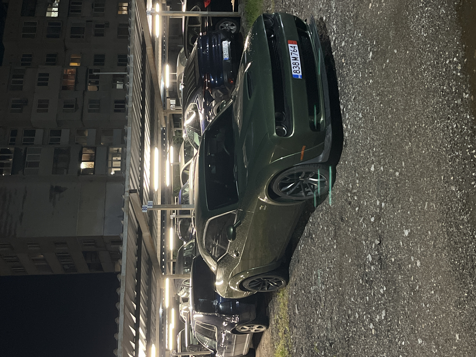 The image depicts a dark green Dodge Challenger parked at night in a covered parking structure. The car is positioned in the foreground, facing towards the viewer's right, and prominently displays a Bulgarian license plate "838 M 764" with the "BG" country code and EU stars emblem. The ground surface appears dark and somewhat damp, possibly due to rain.
Behind the Challenger, several other vehicles are parked under the bright, long fluorescent lights of the parking structure. A dark blue car directly behind the Challenger also has a visible Bulgarian license plate, "CB 0369 TE," confirming the location context of Sofia, Bulgaria. A support beam within the parking area shows the numbers "80" and "81," likely indicating parking space designations.
In the left background, a large, multi-story apartment building is visible against the dark night sky. Many of its windows are illuminated, suggesting residential use and activity within. The overall scene is a quiet urban night, with no people visible and all vehicles stationary.