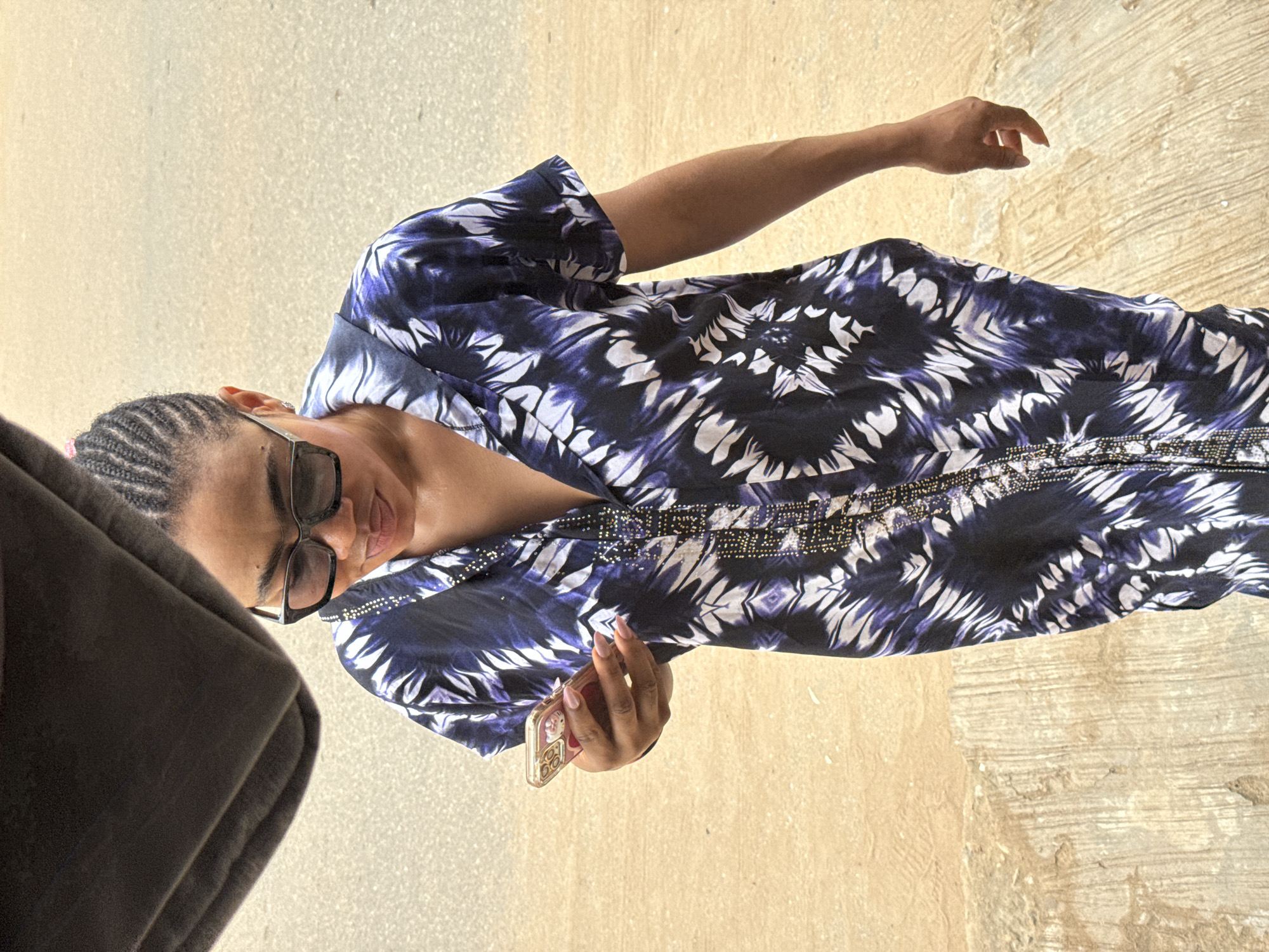 A person wearing a dark blue and white tie-dye dress and sunglasses is looking down at their phone. The person has braided hair. They are standing on a textured, light-colored surface with a darker surface behind them. The scene appears to be outdoors in what could be an urban environment in Abuja, Nigeria, possibly a street or walkway.