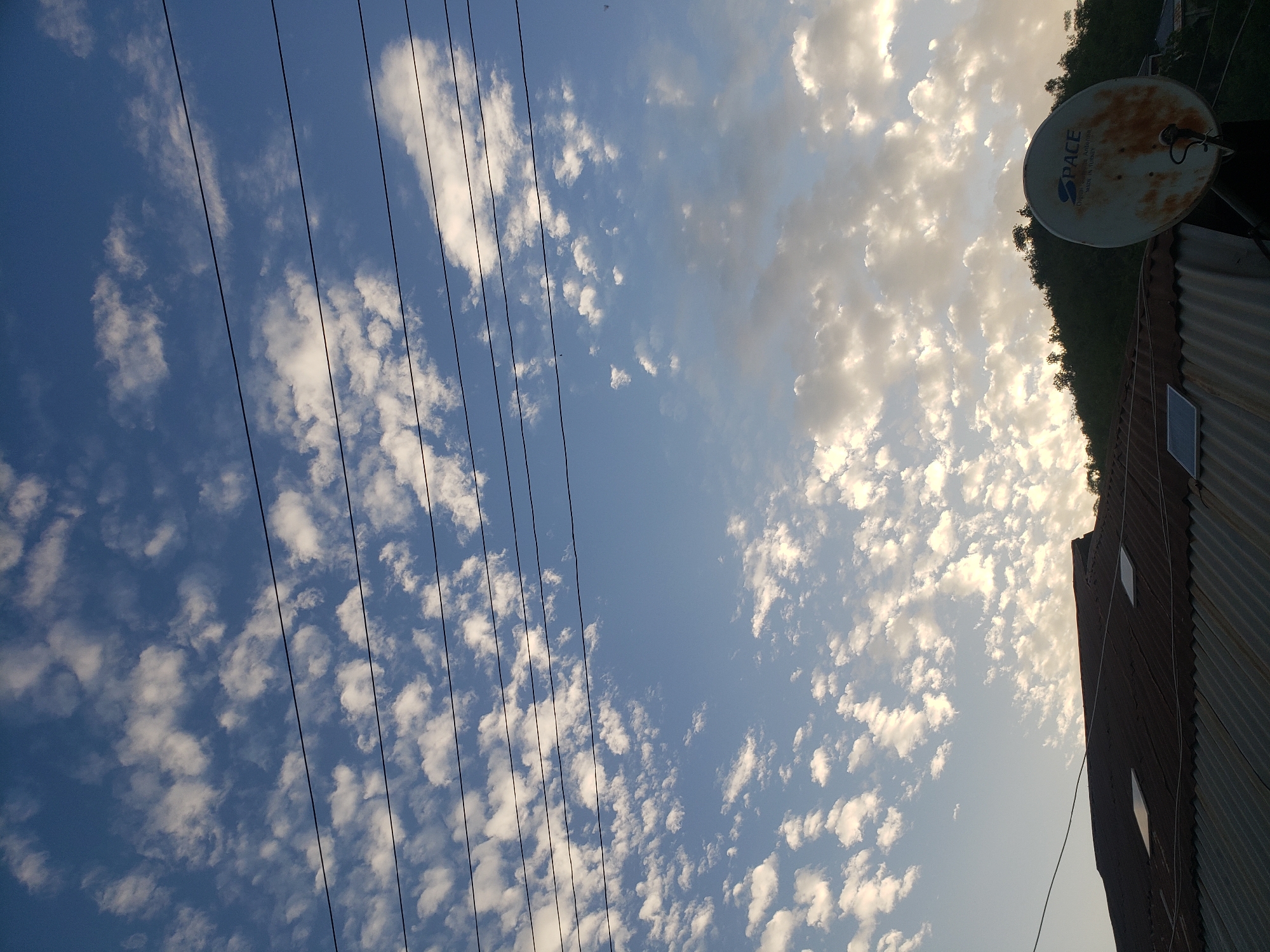 This horizontal image captures a clear blue sky interspersed with scattered white, fluffy clouds. Utility lines or power lines extend across the mid-ground from left to right. The lighting suggests either early morning or late afternoon, with the sun casting a warm glow on some of the clouds on the right side of the frame.
On the far right, part of a building with a corrugated metal roof is visible. An old, slightly rusted white satellite dish is mounted on this roof. Text on the satellite dish clearly reads "PACE," "ORIGINAL QUALITY RECEIVER," and "MADE IN TURKEY." Behind the roof and satellite dish, dense green foliage, likely trees, can be seen, possibly indicating a hillside or natural landscape in the background. A small rectangular panel, potentially a solar panel or a small window, is also visible on the corrugated roof structure below the satellite dish. The scene depicts a tranquil, everyday view from an elevated position, likely a rooftop, in Lokoja, Nigeria.