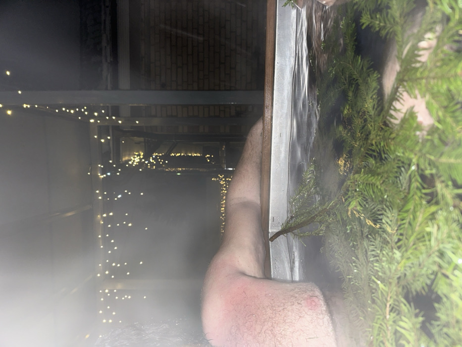 The image depicts an outdoor night scene in Базарово, Russia. A person is partially immersed in a hot tub, with their upper torso and right arm visible above the steaming water. The skin on the visible upper right arm and shoulder exhibits noticeable redness and is covered in hair. The hot tub contains water with steam rising from its surface, and several fresh evergreen branches are floating within it. Overhead and to the right, multiple strands of string lights with warm yellow bulbs provide illumination. A brick building and metal structural elements are discernible in the background.