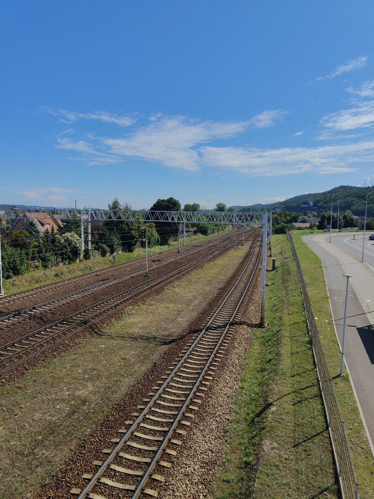 The image captures a view of multiple railway tracks in Rumia, Poland, extending into the distance under a bright blue sky with scattered white clouds. The tracks, laid on ballast, are bordered by grassy areas and a paved road with a sidewalk and a metal fence. On the left side of the tracks, residential buildings with red roofs and lush green trees are visible. In the background, a lush green hillside rises, featuring some structures, possibly industrial or historical, and more dense vegetation. The scene suggests a sunny day in a semi-urban or suburban environment.