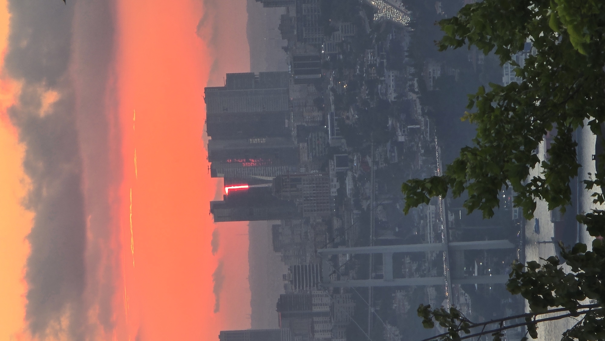An aerial, eye-level, long shot captures a hazy cityscape at sunset in Üsküdar, Turkey, dominated by a vibrant orange and pink sky. In the mid-ground, a collection of tall, modern buildings stands out, with one building featuring a prominent red "1" illuminated on its facade. A large suspension bridge spans the water in the foreground, with traffic visible on its roadway. The water below the bridge is calm, with a few boats present. Lush green foliage occupies the bottom portion of the frame, framing the scene and adding a natural element to the urban landscape. The overall impression is one of a bustling city transitioning into evening.