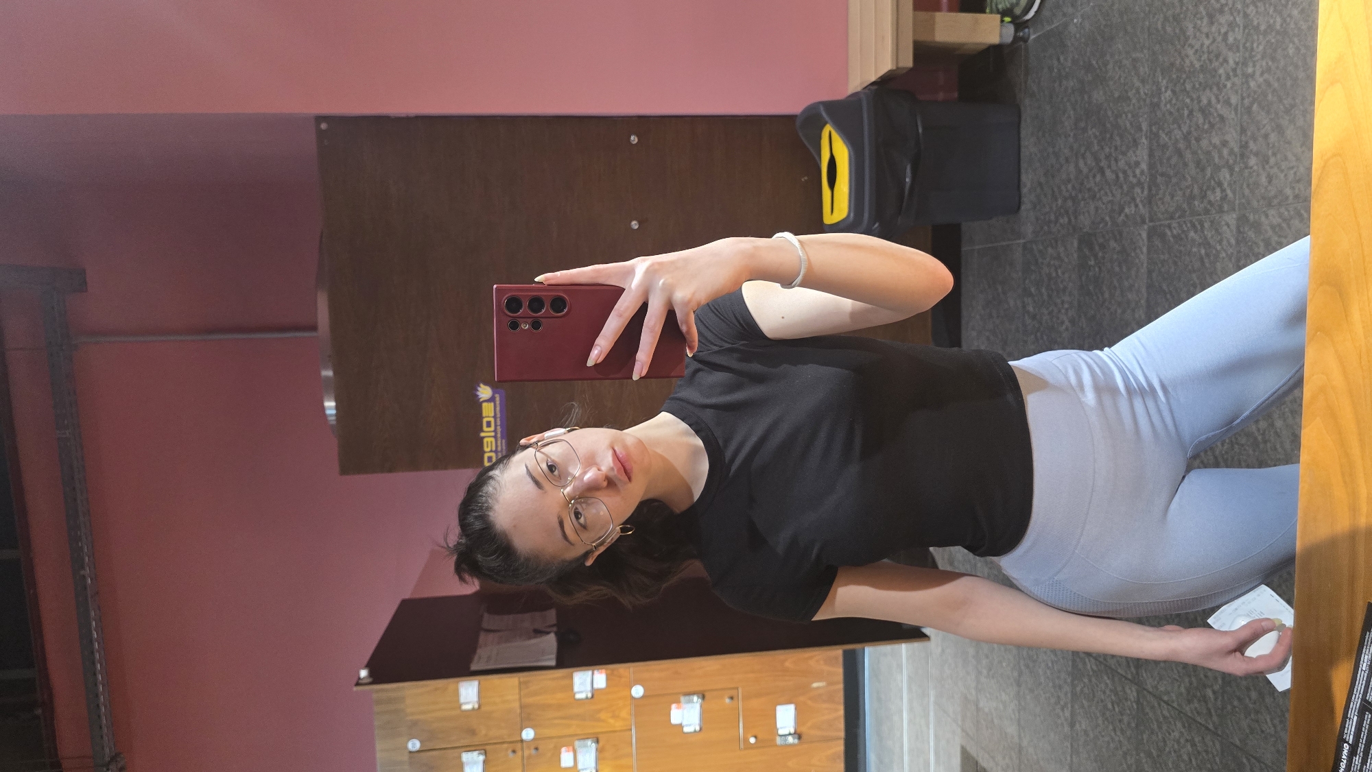 An indoor mirror selfie captures a young woman posing in what appears to be a gym or fitness facility in Stara Zagora, Bulgaria. She is holding a red smartphone with her right hand to take the photo, looking directly at the camera with a subtle smile. She wears a black short-sleeved t-shirt, light blue/grey form-fitting leggings, and gold-rimmed glasses. A silver bracelet is visible on her left wrist. Her dark hair is pulled back, with some strands framing her face.
The background features reddish-pink walls and a dark grey tiled floor. To her left, several brown wooden lockers or storage units are visible, along with a metal shelving unit further back. On the right, part of a black bag with a bright yellow accent is seen. A white piece of paper or receipt lies on the floor near her left hand.
Visible text, appearing reversed in the mirror reflection, includes "G9IQe" and "ONATHM" on a purple sticker with a yellow logo, located on a wooden surface behind her head. Some indistinct text is also present on the white paper on the floor. The setting is well-lit, suggesting an active time of day.