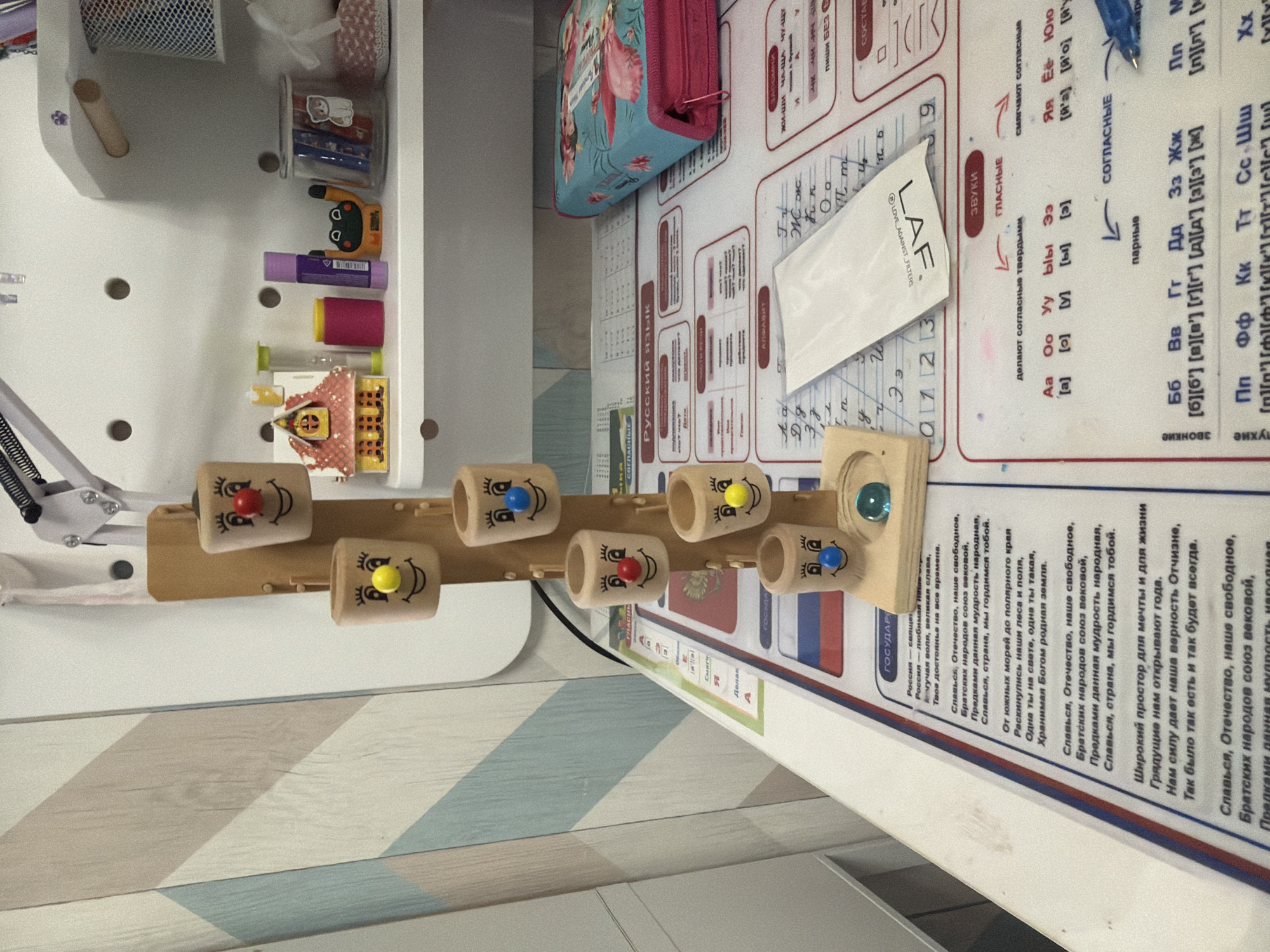 The image shows a desk with various items on it. On the left side of the frame is a white shelf unit with small items inside. There's a pencil case and a white packet. There is a Russian learning mat on the right side with the Russian alphabet, short texts, and the Russian flag displayed. There is a wooden toy with holes and colorful spheres in them. The surface has a chevron pattern.