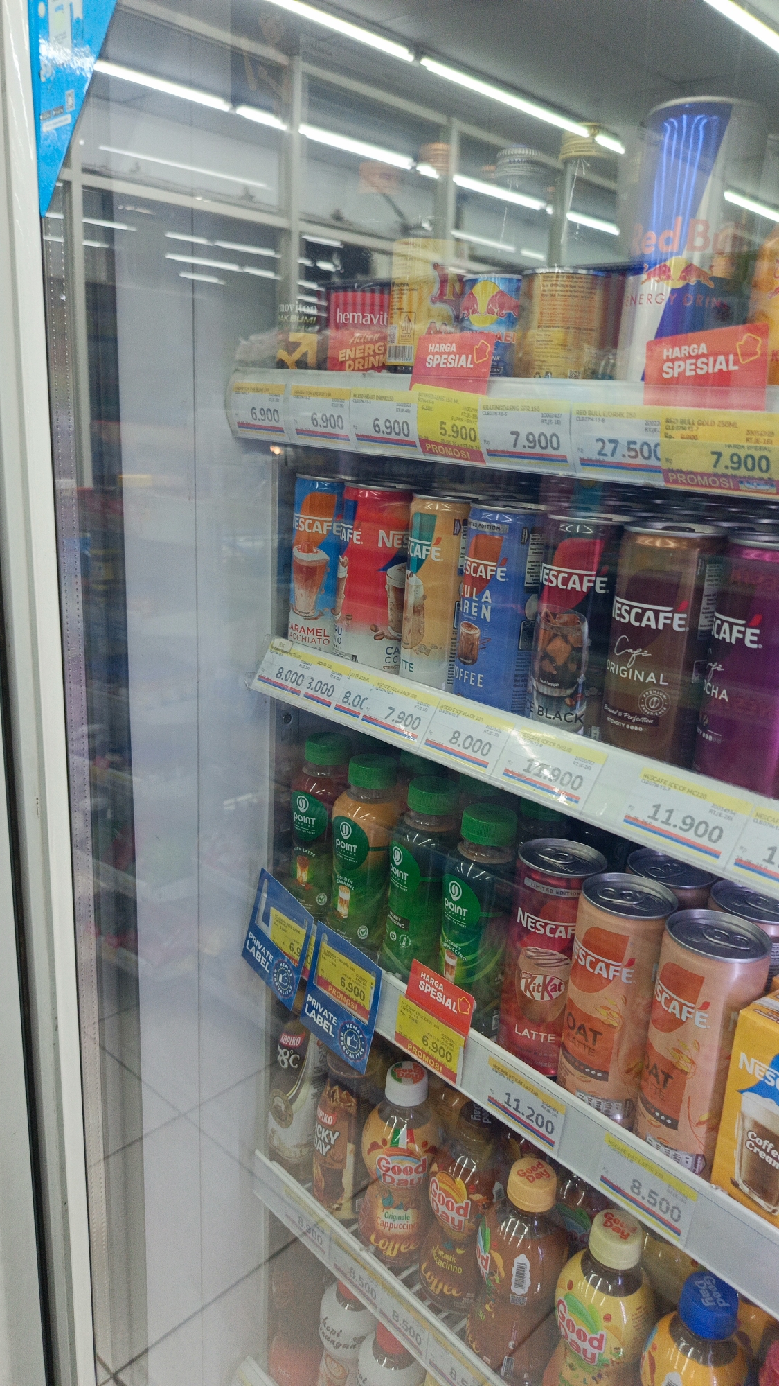 The image shows the interior of a refrigerated display case in a convenience store, likely located in Margajaya, Indonesia, given the context of local product labeling. The display case is filled with various beverages, predominantly canned energy drinks and coffee drinks, as well as some bottled drinks.
On the top shelf, several cans of Red Bull energy drink are visible, along with other brands of canned drinks, some with Indonesian labels like "HEMAVIT." Price tags are displayed below each item, with prices such as "6.900," "5.900," and "27.500." One prominent price tag reads "HARGA SPESIAL," indicating a special offer.
The middle shelf features an assortment of Nescafé canned coffee drinks in various flavors and Nescafé Original cans. There are also some Nescafé coffee bottles. Price tags for these items include "8.000," "7.900," and "11.900." Below these, a shelf displays bottled drinks, mostly from the "point" brand, in green and orange bottles with green caps. Another Nescafé product with a "Coffee Creamy" label is visible on the far right.
The bottom shelf shows more bottled drinks, including "Good Day" brand coffees like "Cappuccino" and "Caramelo Macchiato," and "Good Day" flavored drinks in yellow bottles. Other bottled drinks with white labels are also present. Price tags for this shelf include "8.500."
The overall environment appears to be a retail setting, with the refrigerated case being the main subject. The lighting is consistent with interior store lighting, and reflections from the glass door of the display case are visible. No people are directly depicted within the frame of the display case itself.