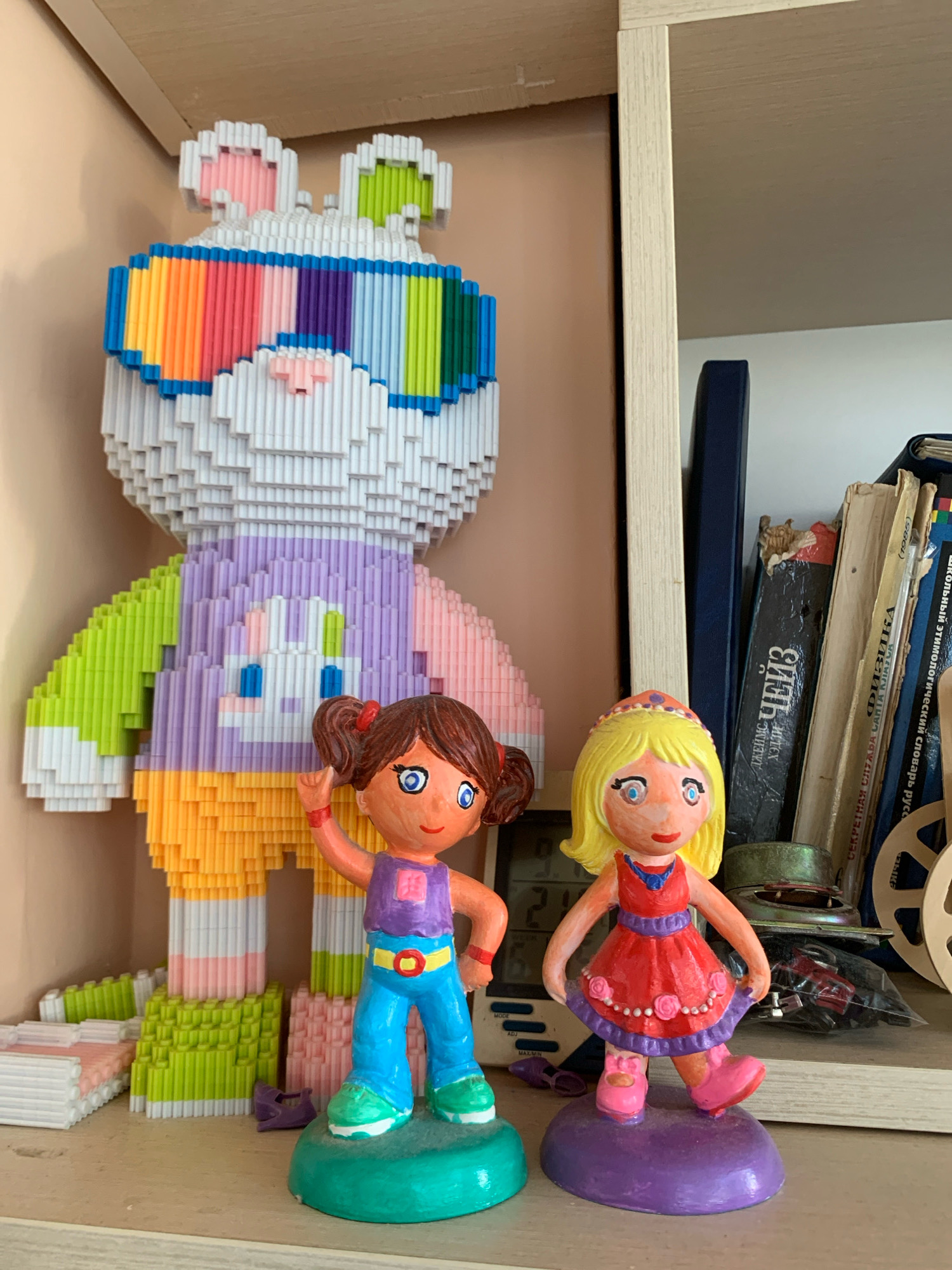 The image shows a shelf with several objects, including a large pixelated bunny figure and two small dolls. The bunny is predominantly white with colorful accents in its sunglasses, vest, and ears. It stands on a pastel-colored base made of interlocking blocks. To the right of the bunny are two small, hand-painted dolls. The doll on the left has brown pigtails and is wearing a purple top and blue pants. The doll on the right has blonde hair and a red dress. Behind the dolls and to the right, there are several books and some electronic components. The shelf appears to be made of wood. The image is taken from a medium distance, with the shelf filled with various items. The lighting suggests an indoor environment, likely a room. The location is a shelf in an apartment in Minsk, Belarus.