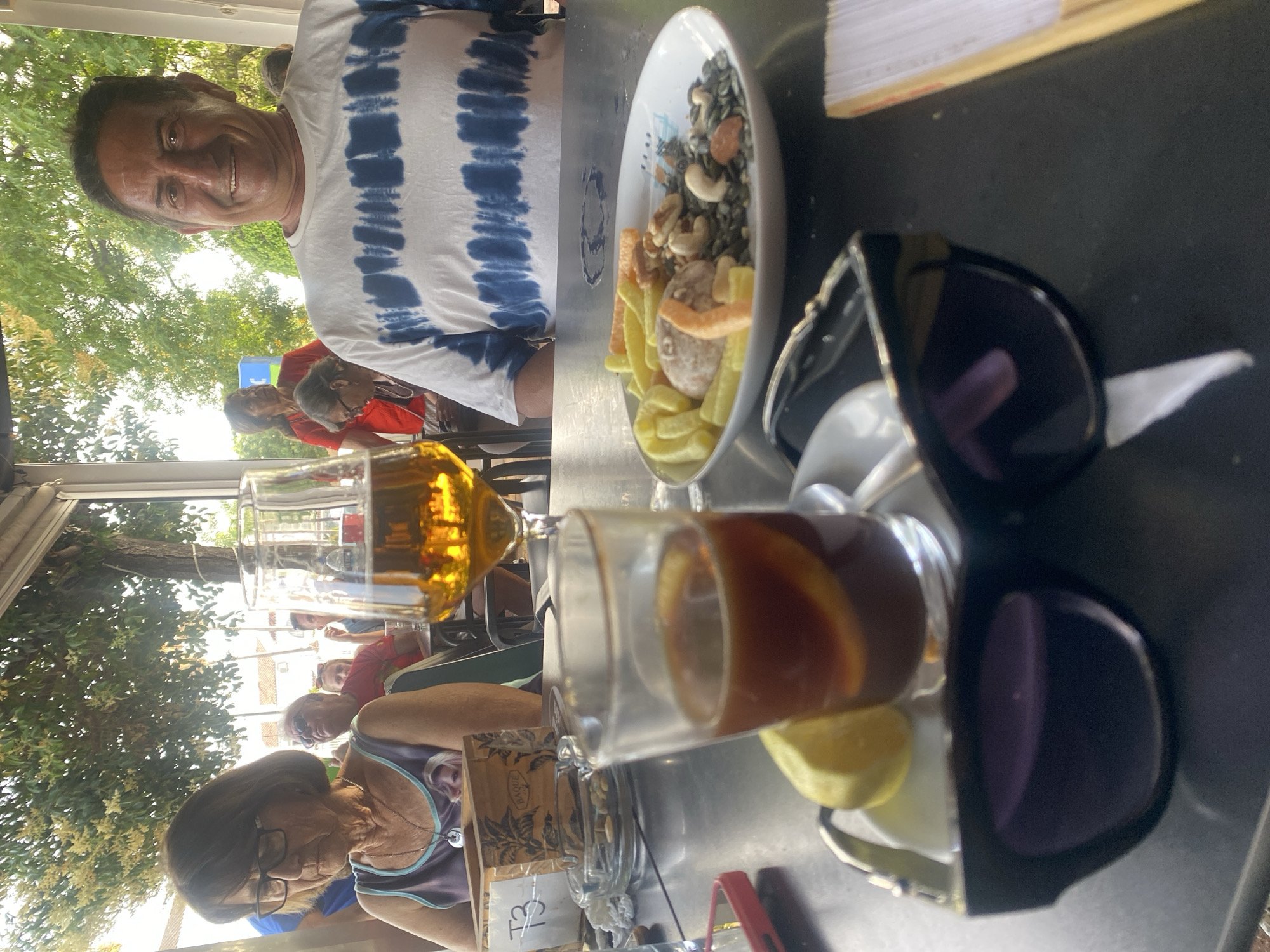 This image was taken at an outdoor cafe in San Pedro De Alcántara, Spain. In the foreground, a pair of black sunglasses rests on a dark table, obscuring a portion of a small white saucer holding a drink with lemon and ice, and a wedge of lemon. To the right of the saucer is a white plate filled with a mix of nuts and snack crackers. A stemmed glass filled with amber liquid, likely beer, is visible to the left of the plate. In the background, a man with dark hair and a blue and white tie-dye t-shirt is smiling at the camera. To his left, a woman with short brown hair and glasses wears a patterned tank top. Other patrons and trees are visible in the background, suggesting a relaxed outdoor dining atmosphere.