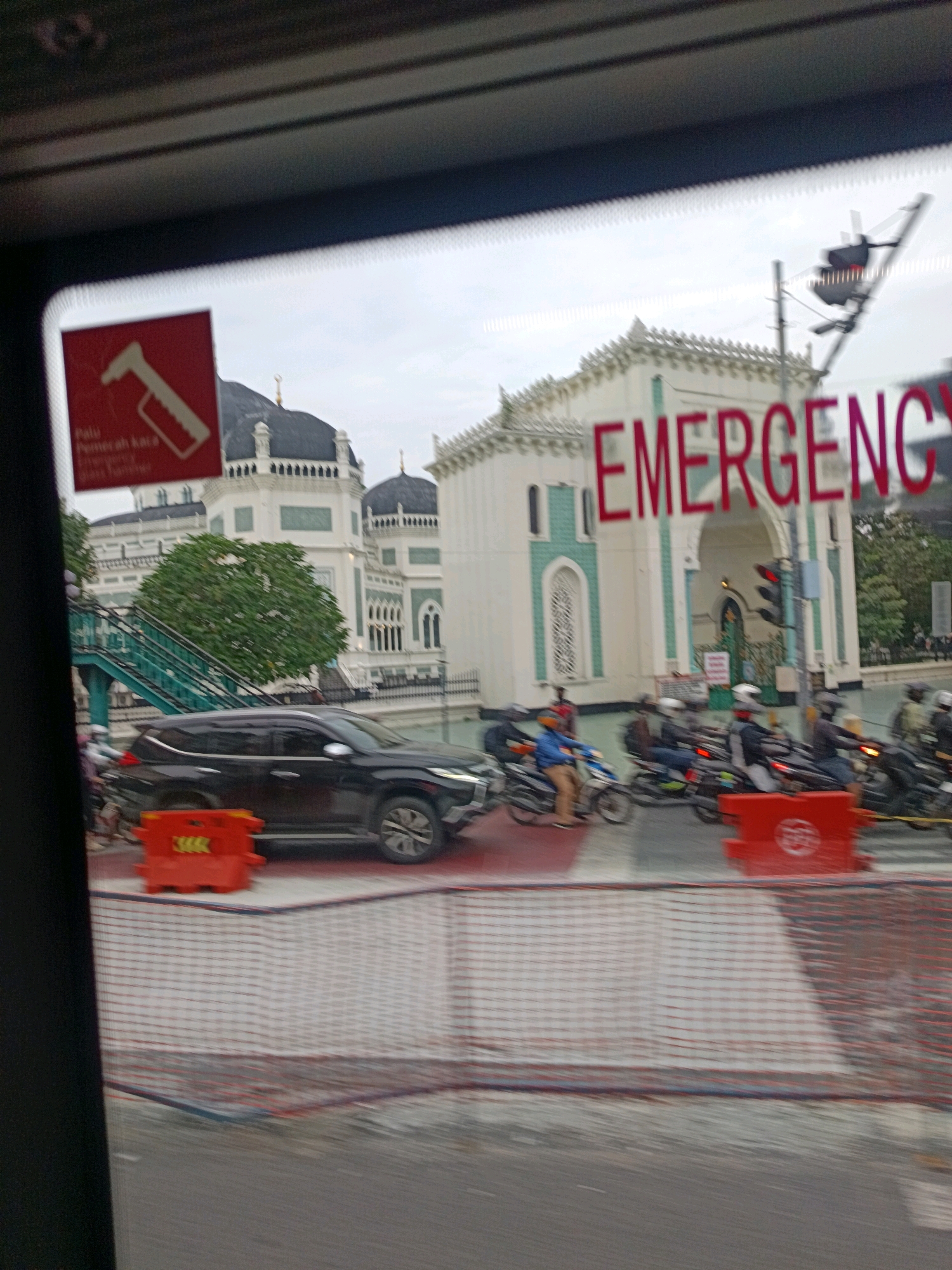 The image captures a view through a vehicle's window in the City of Medan, Indonesia. In the foreground, a red sign with an icon of a hammer and text in Indonesian, reading "Palu Pemecah Kaca Emergency," is visible on the left. Across the street, a large, white building with green accents and multiple domes, resembling a mosque, is prominently featured. The word "EMERGENCY" is written in large red letters across the building's facade. Several motorcycles are seen on the road, with riders wearing helmets, suggesting traffic is present. A black SUV is also parked near the building. Red safety barriers are placed on the road. The environment appears to be a city street with moderate traffic and pedestrian activity.