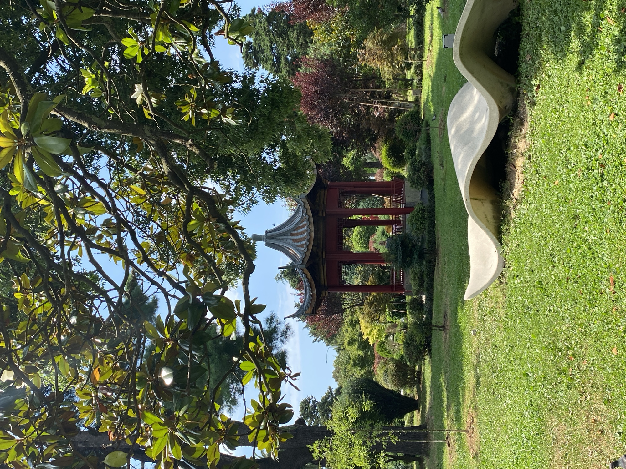 The image depicts a serene park scene with a distinctive red, multi-tiered pagoda structure in the background, partially obscured by lush green trees. In the foreground, a large, abstract, wave-like concrete sculpture rests on a grassy lawn. The park appears to be a well-maintained garden with various types of trees and shrubs, including those with red foliage. The sky is a clear blue with scattered clouds. This scene is located in Batumi, Georgia, at the Batumi Botanical Garden. There are no visible people in the frame. The overall atmosphere is peaceful and idyllic, suggesting a popular spot for relaxation and nature appreciation.