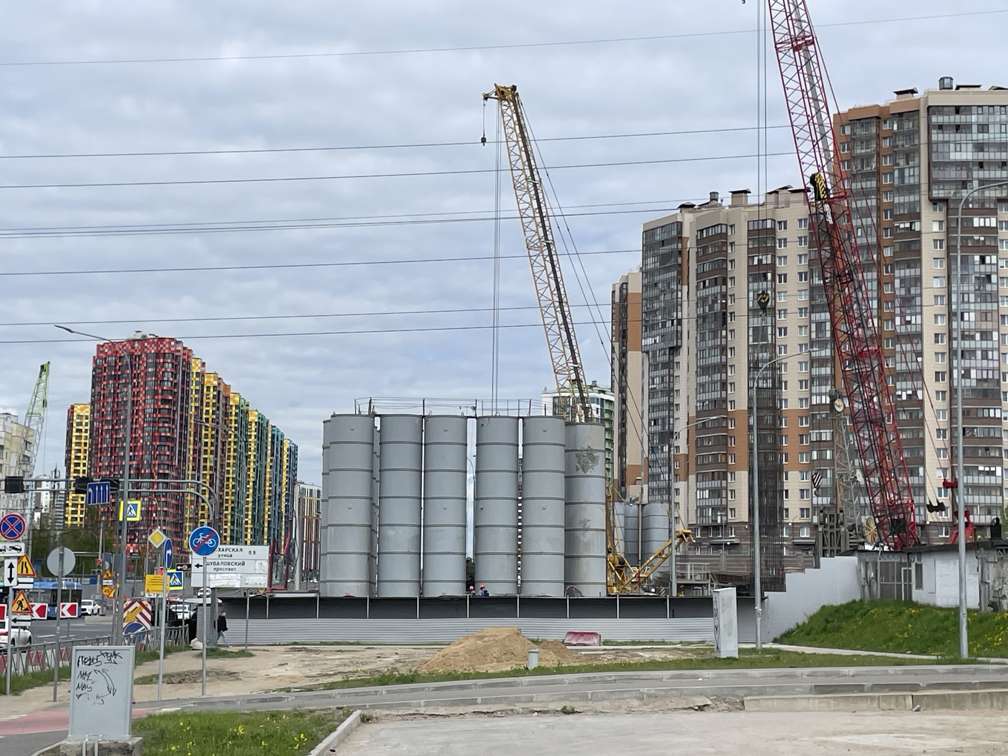 The image shows a construction site in Saint Petersburg, Russia. In the background are high-rise residential buildings, characterized by colorful facades. In the foreground, there are several large, cylindrical, gray storage tanks. Construction cranes are visible in the middle ground, including a yellow crane and a red crane. Street signs and a traffic intersection are situated on the left side of the frame. In the immediate foreground is a paved area with some greenery. There are two people wearing orange safety vests in the middle ground in front of the silos.