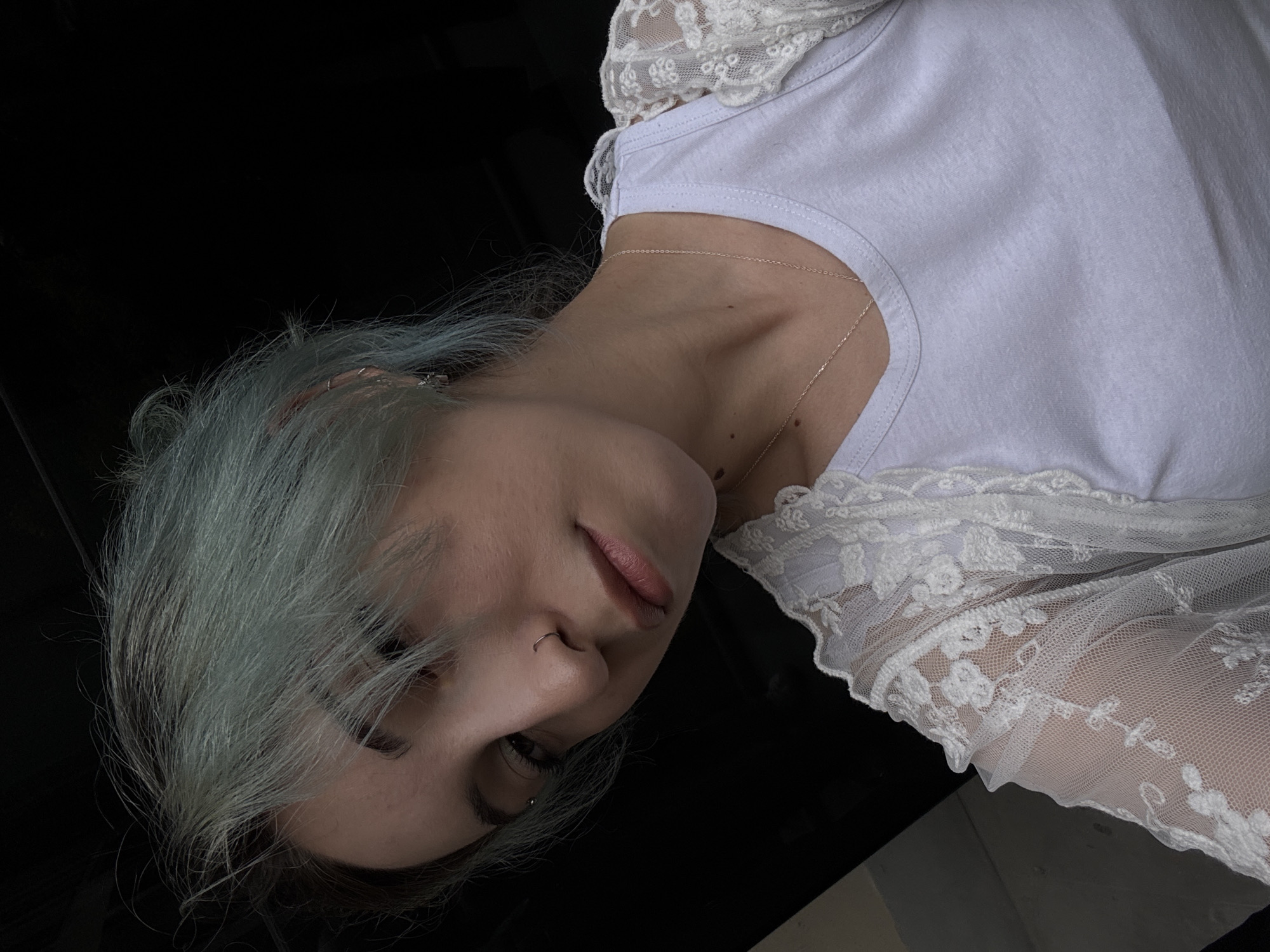 A photograph depicts a young woman with light blue hair wearing a white t-shirt with lace detailing and a delicate gold necklace. She has a nose ring. The background is primarily dark. The image is a close-up shot, tilted at an angle. The location is not discernible from the image, and there is no indication it was taken in Yerevan, Armenia.