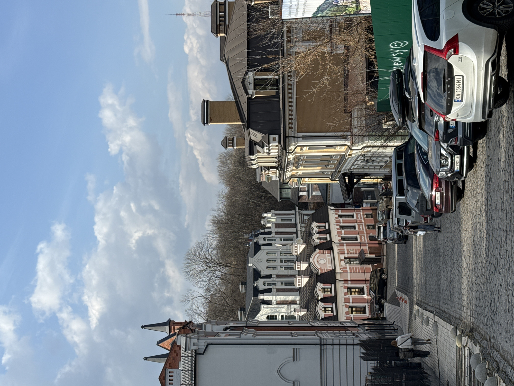 The image depicts Vozdvyzhenska Street in the Podil district of Kyiv, Ukraine, viewed looking downhill on a sunny day with scattered clouds. The cobblestone street slopes distinctly, flanked by a paved sidewalk on the left, segmented by grey concrete bollards.
On the left, a multi-story building with a white facade, dark grey trim, and two prominent black spires on its roofline is visible, partially obscured by a dark metal fence. Further down on the left, a two-story building with a light pink facade, white accents, and a dark grey mansard roof with dormer windows is present. On the opposite side of the street, a multi-story building with a yellow facade, white trim, and a dark grey mansard roof with chimneys is prominent. In the background, beyond these structures, a large white multi-story building with dark brick detailing and a dark grey roof stands atop a rise. A densely wooded hillside with bare deciduous trees forms the distant background, with a slender communication tower visible on the far right horizon.
Three individuals are present: two are walking downhill on the left sidewalk, one in dark clothing and another in a light-colored jacket and jeans. A third individual, wearing a light-colored coat and carrying a bag, walks downhill on the cobblestone street on the right.
Multiple vehicles are parked along the right side of the street, facing downhill. The closest vehicle is a white Volvo V60 T5 AWD station wagon with Ukrainian license plate "KA 1564 MI". Behind it, a dark SUV with a black roof cargo box, a dark Nissan SUV with Ukrainian license plate "AP 8939 IE", and several other parked sedans and SUVs are visible. A black SUV is also parked on the left side of the street near the pink building. Two red and white traffic cones are positioned on the left side of the street. A green construction barrier or fence, partially covered in white graffiti, runs along the right side of the street beyond the parked cars, with a large, partially visible billboard or banner featuring a landscape image behind it.