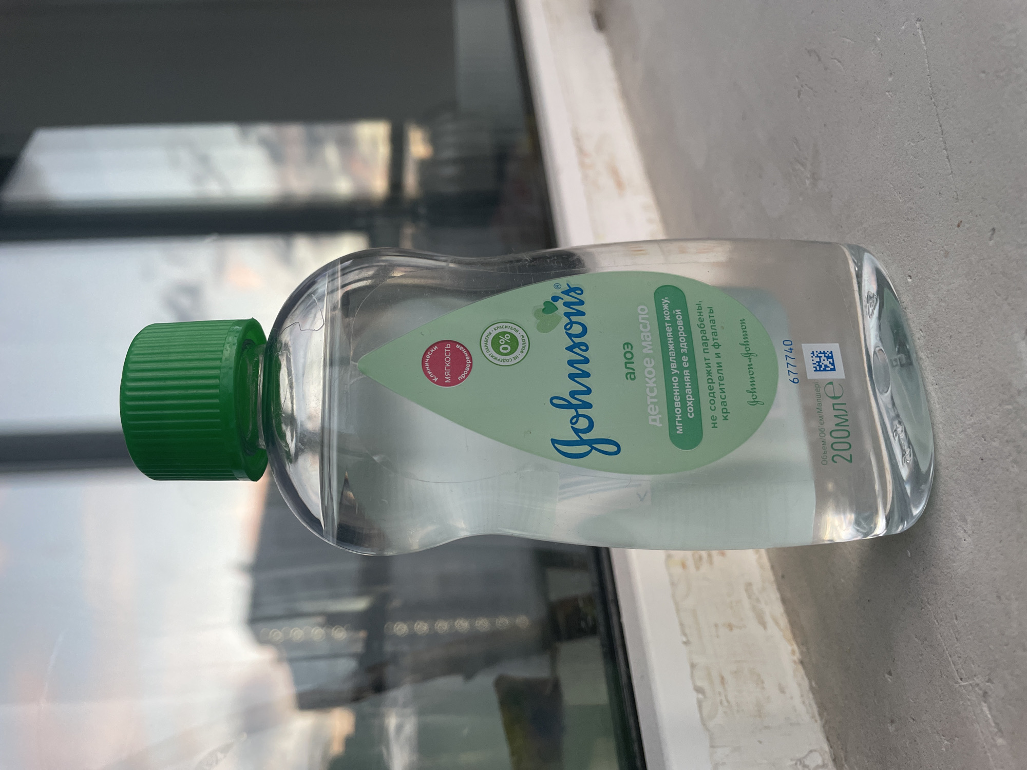 A clear plastic bottle of Johnson's Baby Oil with Aloe is placed on a windowsill. The bottle has a green cap and a label with text in Russian. The background shows a blurred reflection of buildings and a sky with soft colors, suggesting it might be dusk or dawn. The windowsill appears to be made of concrete or stone and is slightly dusty.