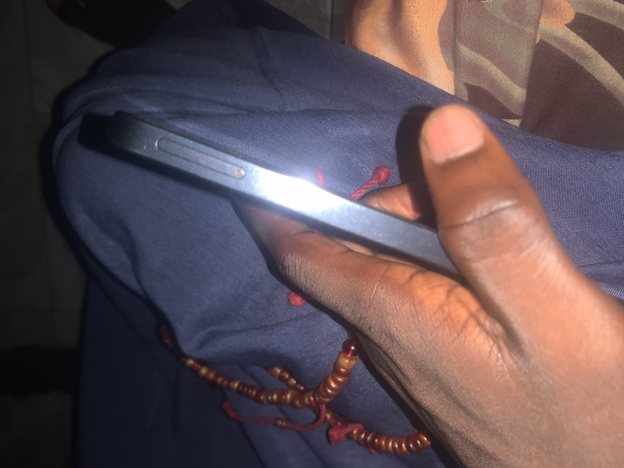 In Jalingo, Nigeria, a close-up shot focuses on a person's hand, holding a smartphone. The phone's metallic edge is visible, reflecting light, and rests against a dark blue fabric. A string of prayer beads, primarily red and brown with some translucent yellow beads, drapes over the fabric near the hand. The lighting is dim, suggesting an indoor or nighttime setting. The background is blurred, with hints of patterned fabric visible to the right.