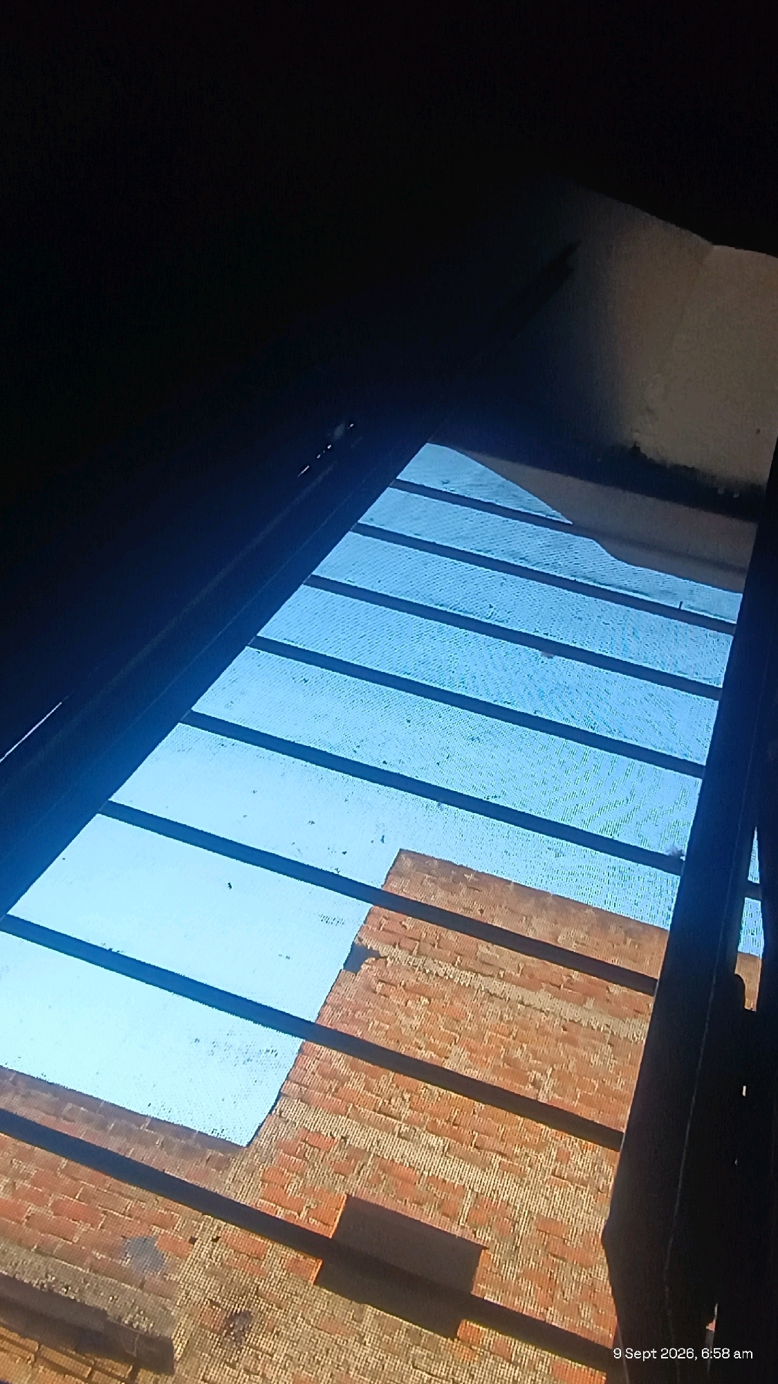 The image shows a low-angle view of a partially covered outdoor area, likely a patio or balcony, with a brick wall in the background. The sky is visible through a slatted covering, which appears to be made of metal bars and a mesh material. Some debris is scattered on the mesh. The brick wall is weathered and shows signs of age. The bottom right corner of the image contains a timestamp: "9 Sept 2026, 6:58 am". The scene is likely located in Udaipur, India, given the context provided. There are no people visible in the image. The overall impression is of an ordinary outdoor space under daylight.