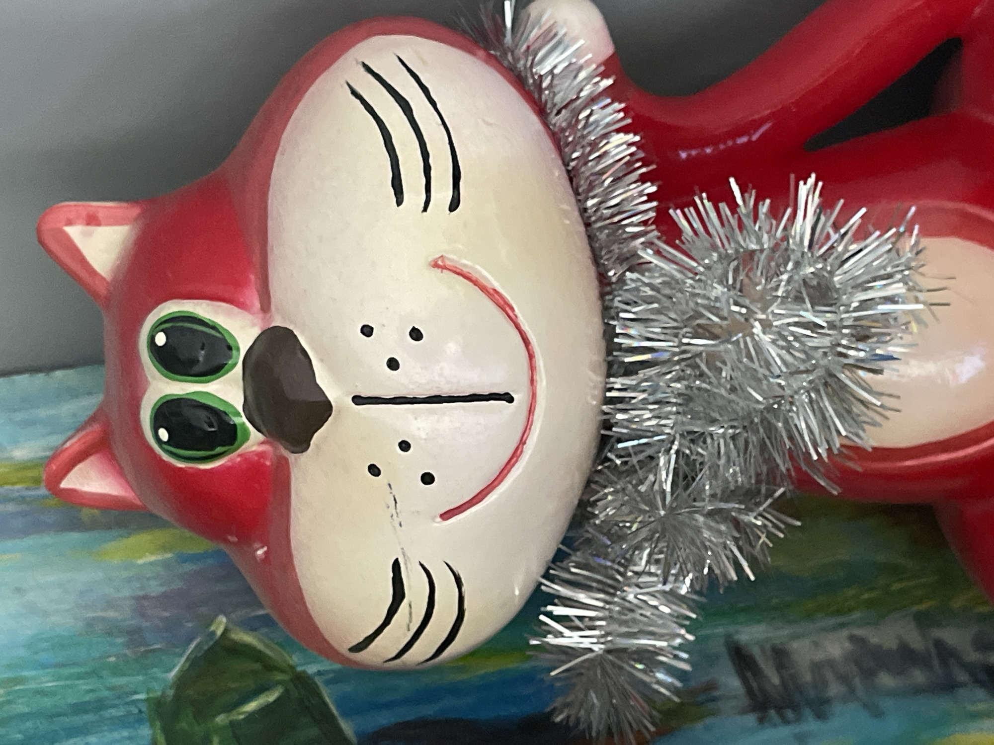 This is a close-up, eye-level, medium shot of a red ceramic cat figurine with white accents. The cat has green eyes with black pupils, a brown triangular nose, and a red smiling mouth with black dots for cheeks and black lines for whiskers. It is wearing a silver tinsel garland around its neck. The background is blurred, showing abstract patterns in shades of blue, green, and yellow, with a hint of gray on the right. The figurine is positioned indoors, with no discernible location in the city of Средний, Russia.
