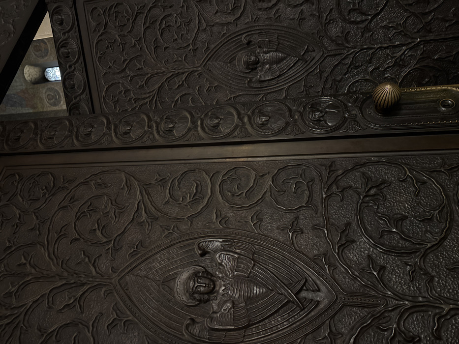 The image depicts a close-up view of the ornate bronze entrance doors of the Alexander Nevsky Cathedral in Sofia, Bulgaria. The doors are dark bronze, intricately embossed with a dense, organic pattern of intertwining vines and stylized leaves across their surface. The visible left door panel features a large, central relief sculpture of an angel with a halo and prominent wings, clad in a tunic, enclosed within a tall, pointed, almond-shaped frame with radiating lines. The right door panel, partially open, displays a similar but smaller angel relief within an identical frame. A narrow vertical bronze pilaster separates the two door panels, adorned with a stack of circular medallions, each containing a bust-length relief portrait of a religious figure. A similar vertical row of medallions is visible along the outer edge of the right door. A prominent, fluted, shell-shaped bronze door handle with an ornate backplate is attached to the lower right door panel. Through the gap at the top of the right door, a dark interior space is visible, revealing an arched window with light-colored glass and fragments of painted frescoes on the wall, depicting circular portraits of additional religious figures. The lighting is subdued.