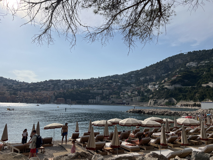 A sun-drenched beach scene is dominated by rows of lounge chairs under striped umbrellas. People relax and stroll along the shore, with a few venturing into the calm water. In the background, a densely populated hillside stretches along the coastline, dotted with buildings nestled among lush greenery. A few boats are visible on the sea. Bare tree branches frame the top of the image, silhouetted against the bright sky.