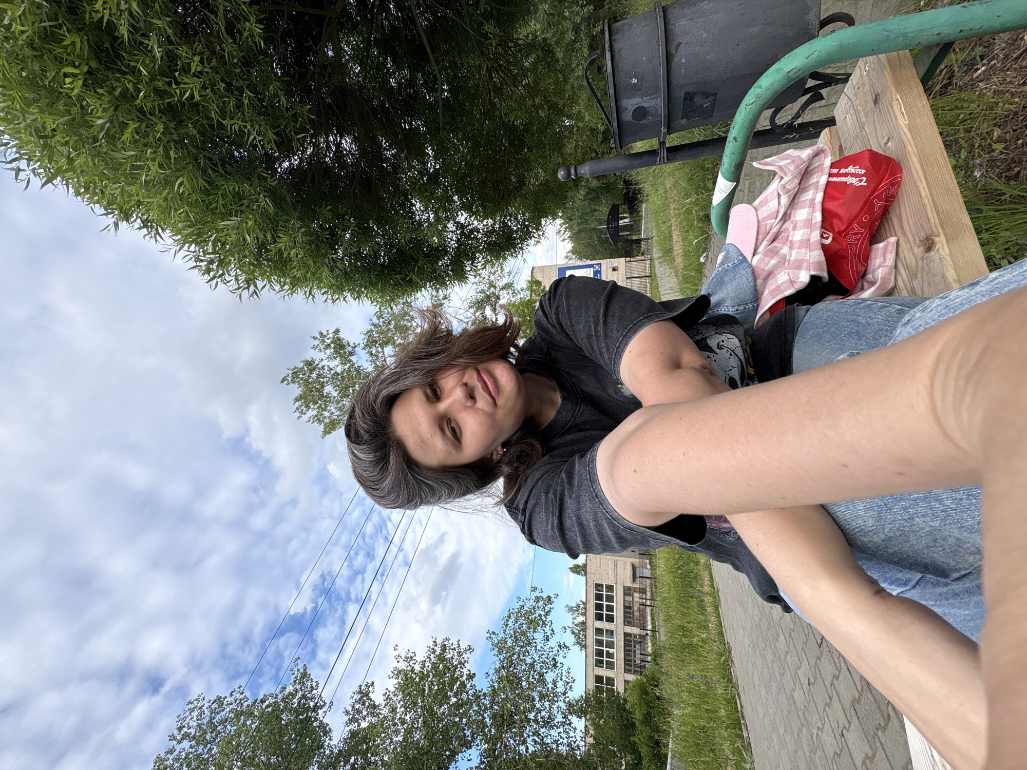 A person is sitting on a park bench, taking a selfie. They have long, dark brown hair with streaks of gray and are wearing a dark t-shirt and blue jeans. Their left arm is resting on their leg, and their right arm is extended to take the photo. On the bench next to them are a pink and white checkered shirt and a red bandana. The background shows lush green trees, a cloudy sky, and a brick building in the distance. The scene appears to be in a park or outdoor public space, possibly in Chaikovsky, Russia. The lighting suggests it is daytime, and the cloudy sky indicates potentially overcast weather. The person's expression is relaxed. Visible text on the red bandana appears to say "Easy Go".