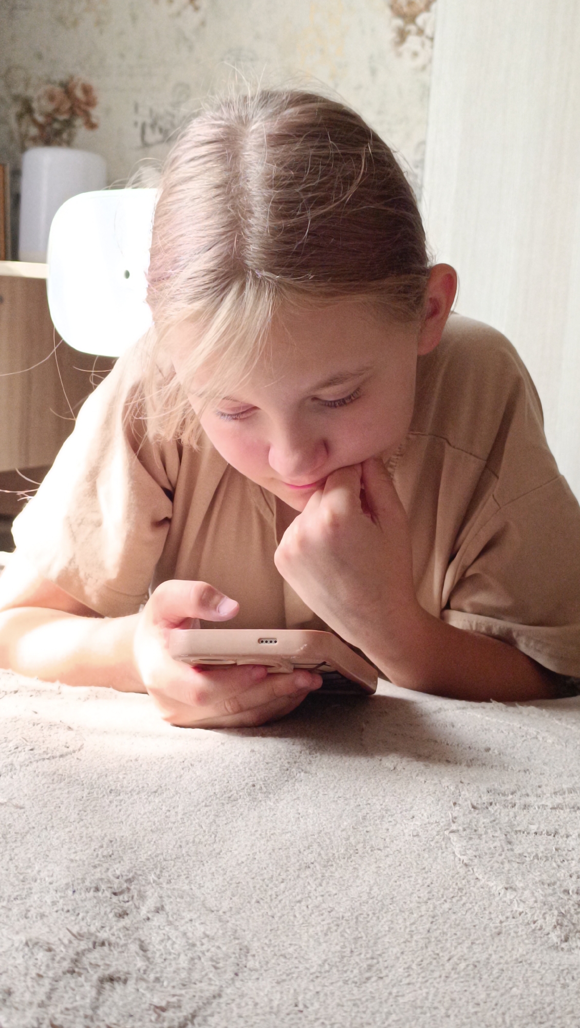 A young girl with blonde hair is looking down at her phone, resting her chin on her hand. She is wearing a light brown t-shirt and is lying on a light-colored, textured surface, possibly a carpet or rug. The background is slightly out of focus, showing a wallpaper with a floral pattern and a white object that appears to be a lamp or a diffuser. The lighting suggests an indoor setting, likely a home. There are no discernible landmarks or features that would indicate a specific location within the city of Tolyatti, Russia.
