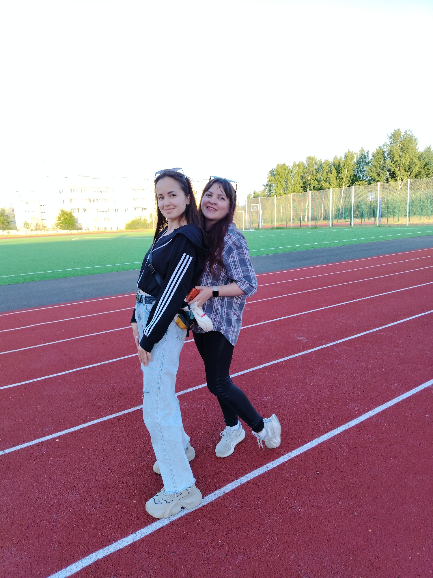 Two women are posing on an outdoor running track in what appears to be a sports complex in Noviy, Russia. The track is red with white lane lines, and behind it is a green artificial turf sports field. Further in the background, there are residential buildings and a line of trees behind a wire fence, indicating an urban or suburban setting.
The woman on the left has long dark hair, wearing a black hooded zip-up jacket with white stripes on the sleeves, light-wash ripped jeans, and chunky light-colored sneakers. She has sunglasses perched on her head and a small nose piercing. A dark cross-body bag is visible across her chest. She is smiling and looking directly at the camera.
The woman on the right, also with long dark hair, is wearing a blue, grey, and white plaid shirt, black leggings, and light-colored sneakers. She also has sunglasses on her head and a dark smartwatch on her left wrist. She is holding a white object, possibly a phone, and a small, colorful packet in her right hand. She is smiling and looking towards the camera, positioned slightly behind the first woman.
The lighting suggests late afternoon or early evening, with a bright sky. Both women appear relaxed and happy. No discernible text is visible in the image.