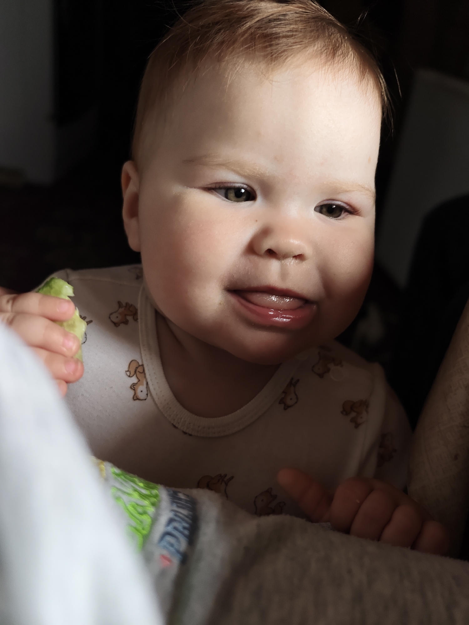 A baby with light brown hair and eyes is visible in a close-up indoor shot. The baby is wearing a white onesie with small brown animal patterns. The baby is holding a piece of green food in their left hand and has their tongue sticking out. The scene appears to be partially lit, with shadows surrounding the baby. The location information is not discernible from the image.