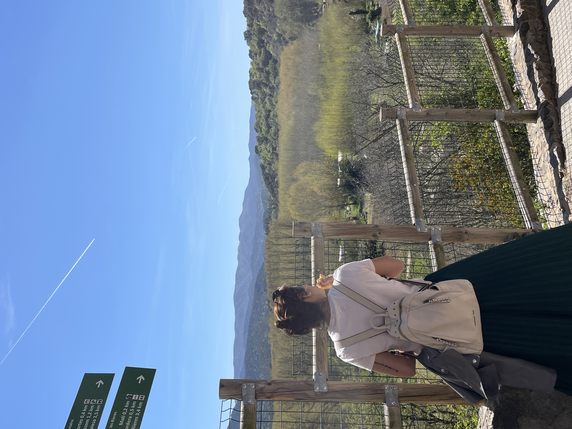 Here is a description of the image:
A woman stands on an elevated platform overlooking a valley in what appears to be the vicinity of Hostalric, Spain. She is leaning against a wooden and wire mesh fence, with her back to the camera, observing the landscape. She has short brown hair and is wearing a white t-shirt, a dark green pleated skirt, and a white backpack. A grey jacket or sweater is slung over her shoulder. She appears to be eating something held to her mouth with her right hand.
The backdrop consists of mountains and a valley with trees. A clear blue sky is visible above, marked by a contrail from an airplane. In the foreground, there are green directional signs providing distances to places such as "Hostalric", "Oortes", "Ell Som", "Collses Altes" and "Molí", "Ordera", "Rieres". The signs are positioned in the lower left corner of the image.
The scene is taken from a slightly angled perspective, with the horizon running diagonally across the frame. The light suggests it is daytime, and the overall impression is that of a tourist enjoying a scenic view.
